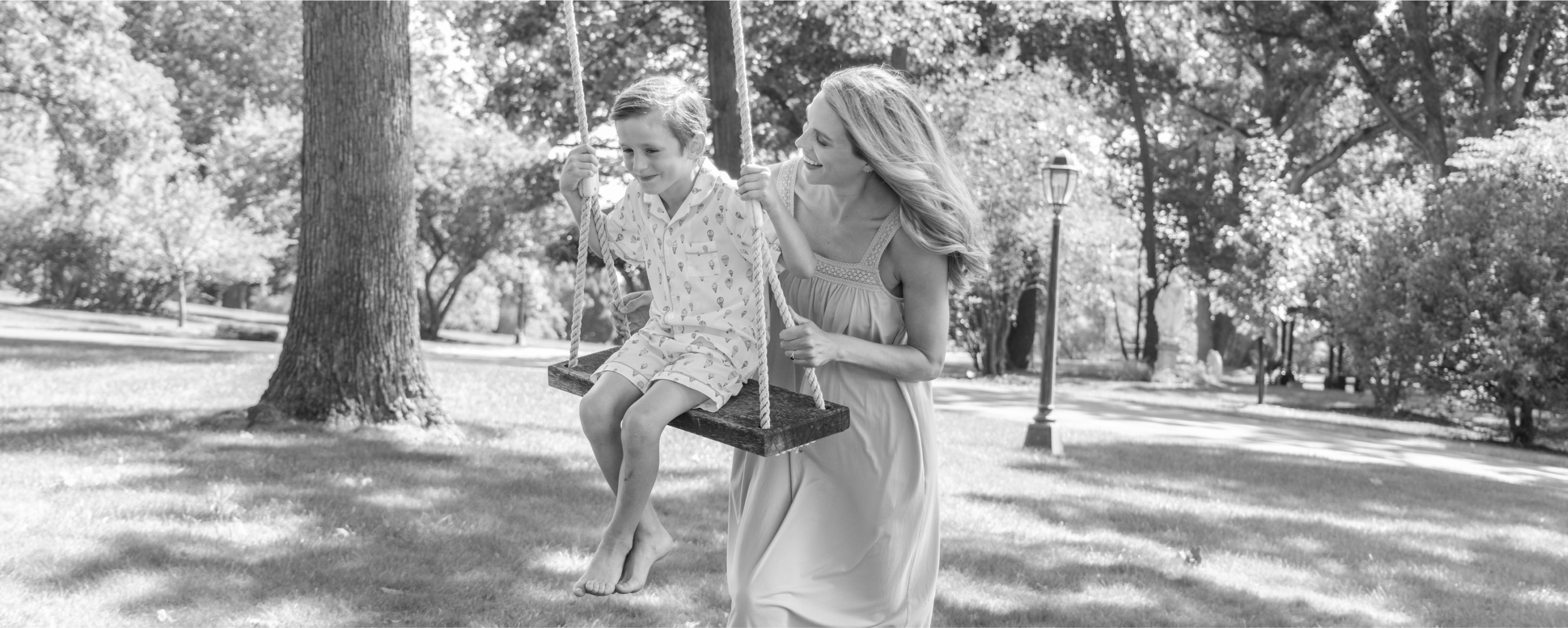 Black and White photo of a Mother and Child outside. The Mother is pushing the child on swing under a tree