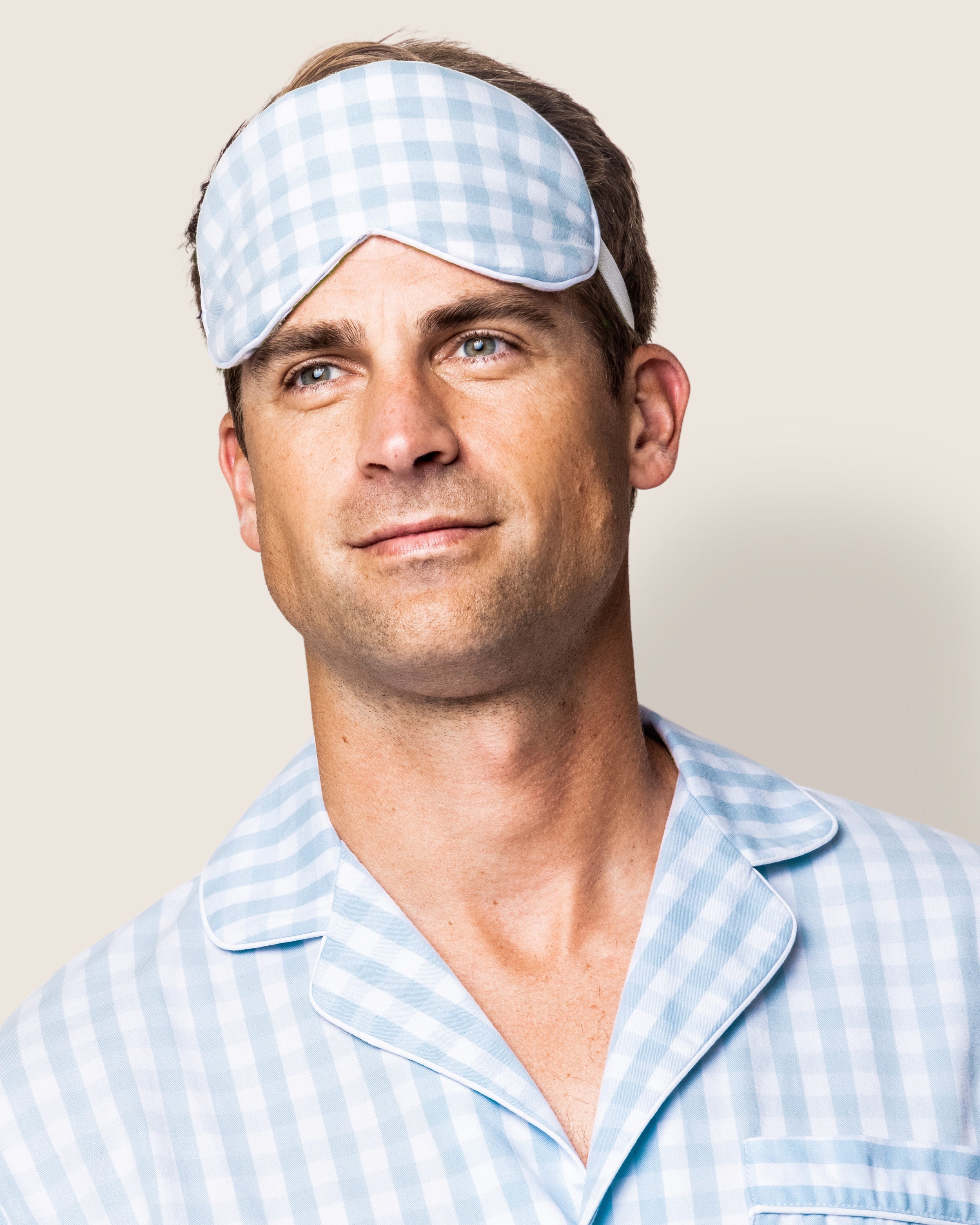 A person stands against a neutral background, wearing a light blue and white checkered pajama top and the Petite Plume Adults Twill Sleep Mask in Light Blue Gingham, looking slightly off to the side with a relaxed expression.