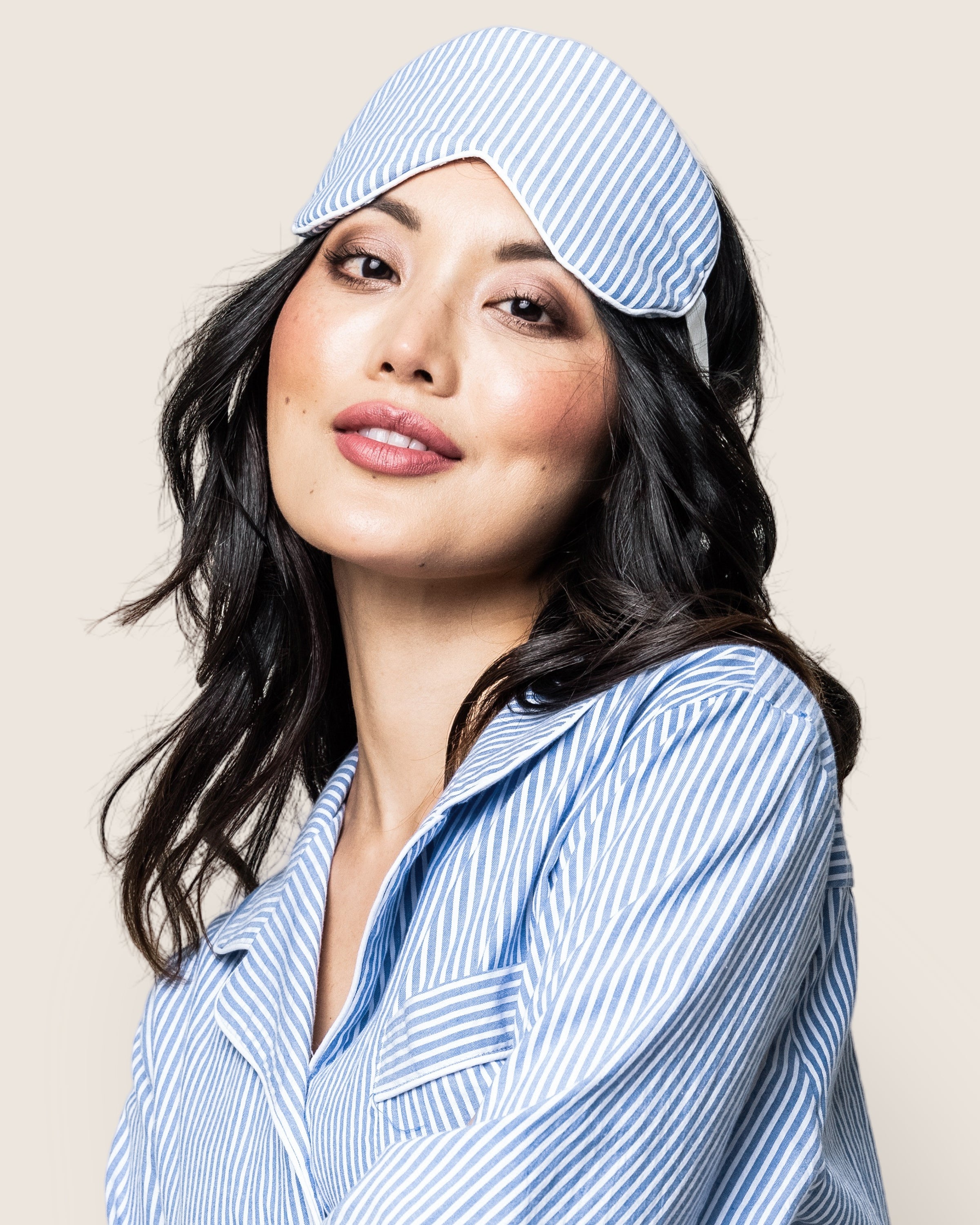 An individual with long dark hair wears Petite Plumes Adults Sleep Mask in French Blue Seersucker atop their head, complemented by matching blue and white striped pajamas. Their neutral expression exudes luxury sleep against a light background.