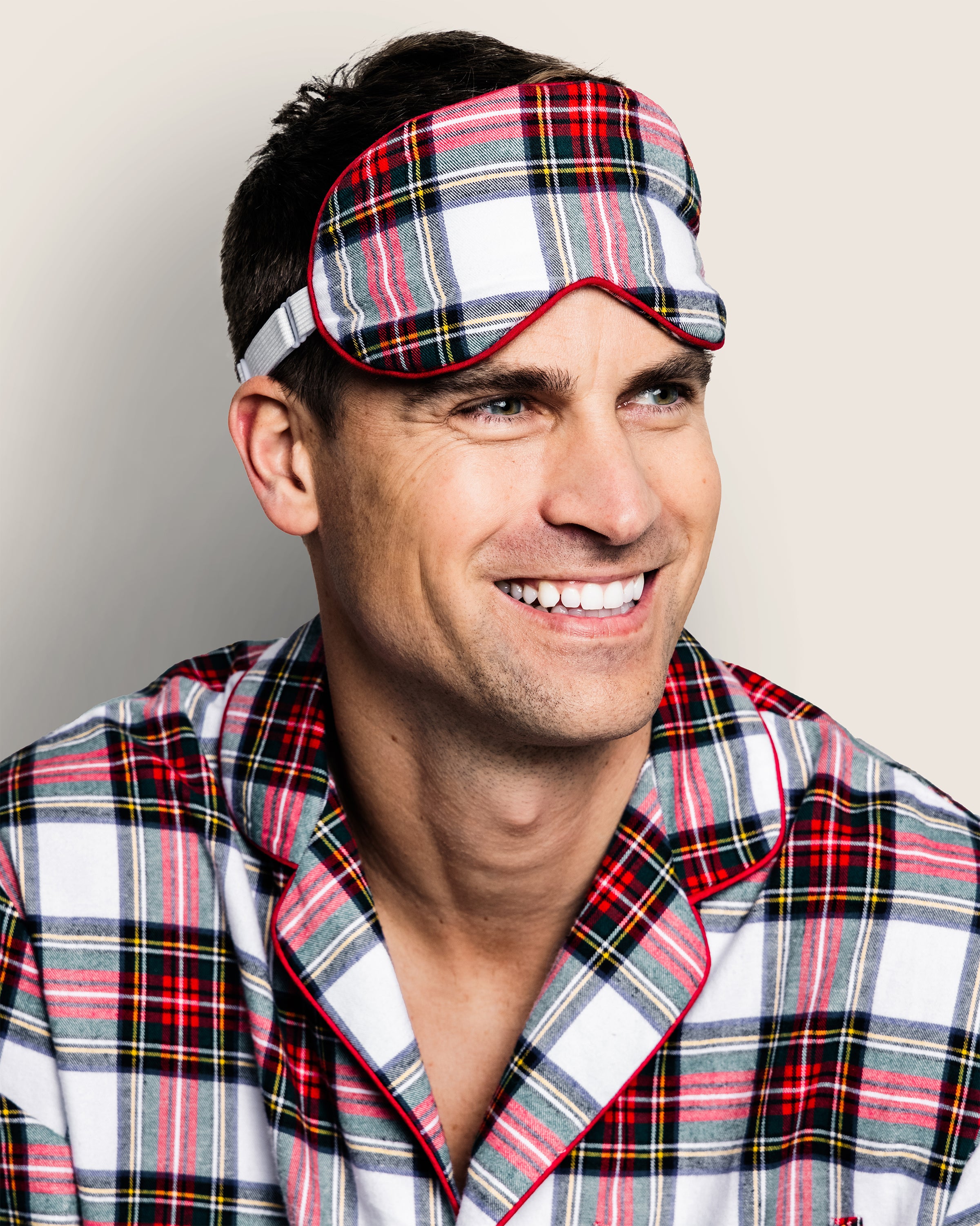 A man smiles while wearing Petite Plumes Adults Brushed Cotton Sleep Mask in Balmoral Tartan, perfectly paired with matching pajamas—an ideal holiday gift.