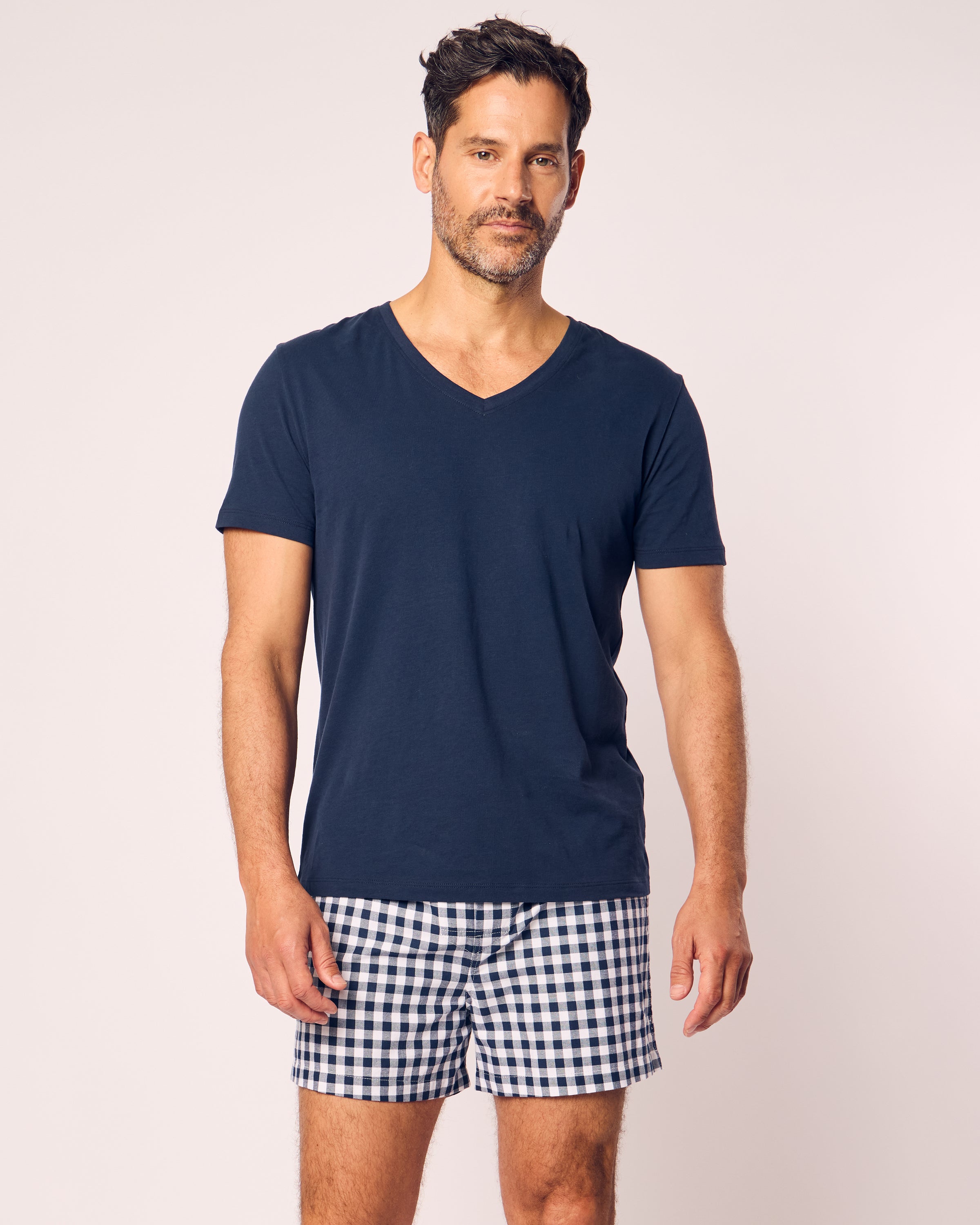 A person in a navy V-neck T-shirt and Petite Plumes black-and-white checkered shorts from the Mens Twill 3 Pack Boxers in Navy stands against a light backdrop. They have short dark hair and a beard.