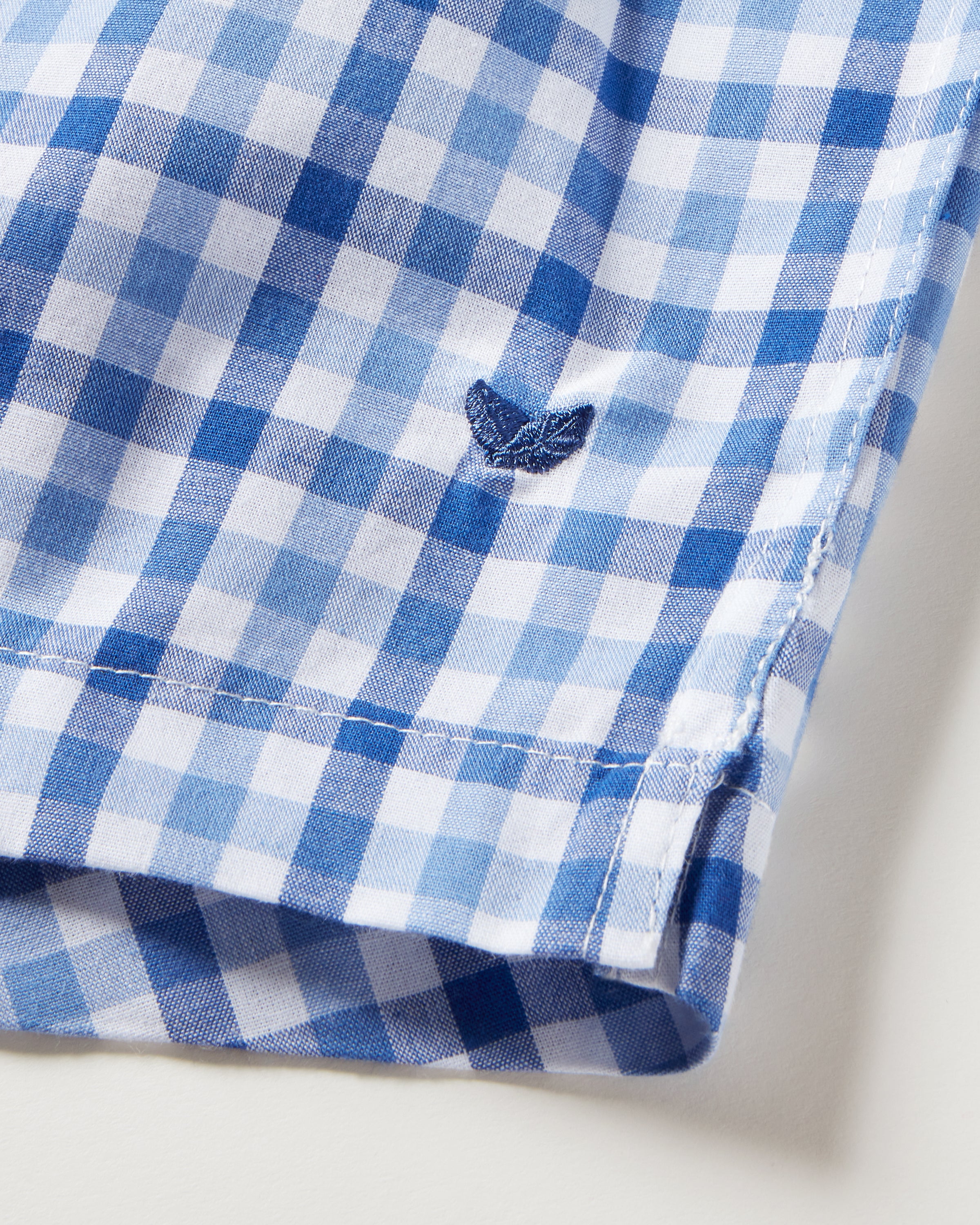 Close-up of a French Blue Seersucker fabric featuring a small embroidered leaf-like logo near the hem. The pattern showcases classic charm with varying shades of blue squares in a gingham style, similar to the Mens Twill 3 Pack Boxers in Royal Blue Gingham by Petite Plume.