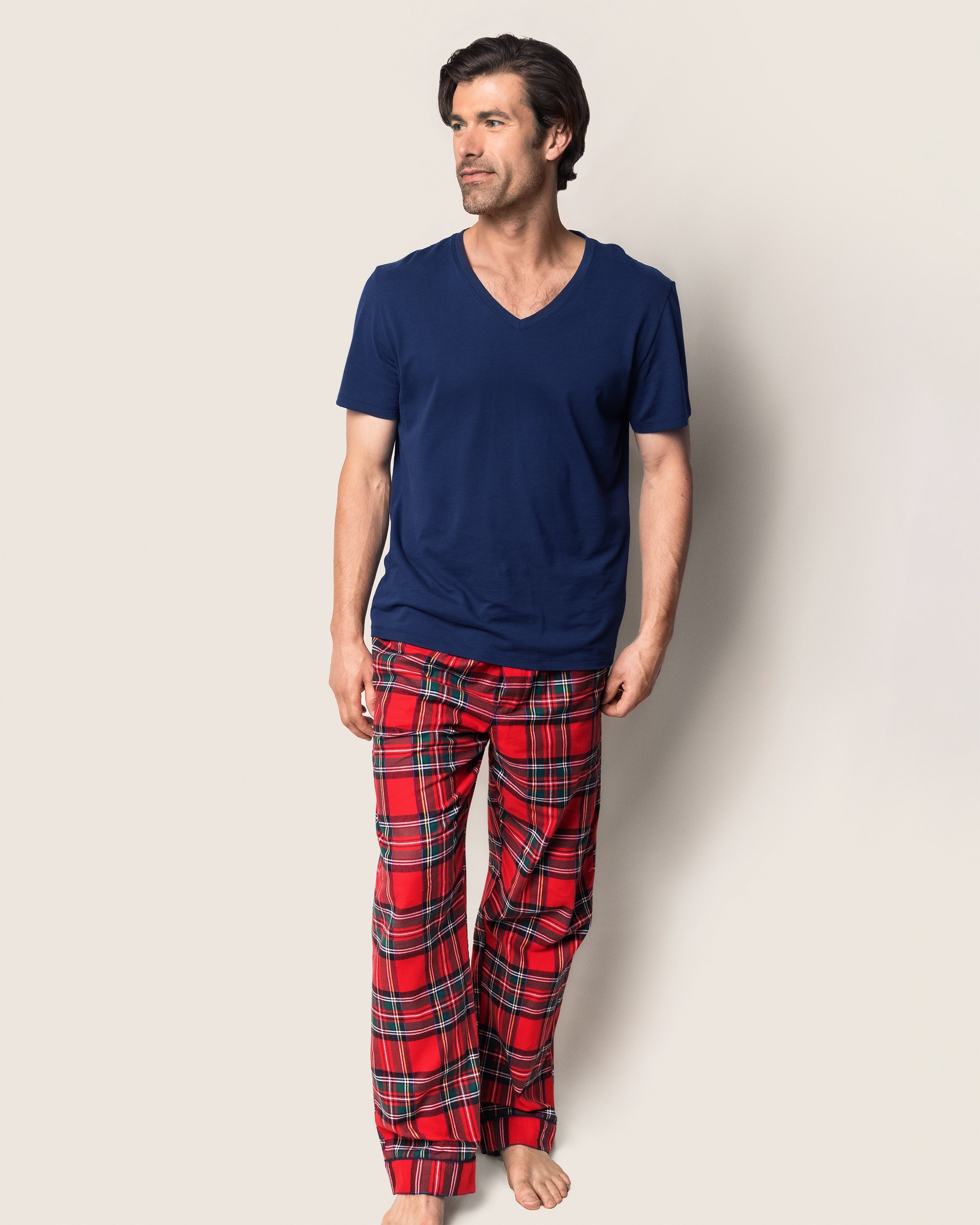 A man stands barefoot wearing a navy V-neck T-shirt and Petite Plumes Mens Brushed Cotton Pants in Imperial Tartan. His hands are relaxed at his sides as he looks left, with the light gray background complementing his luxury sleepwear ensemble.