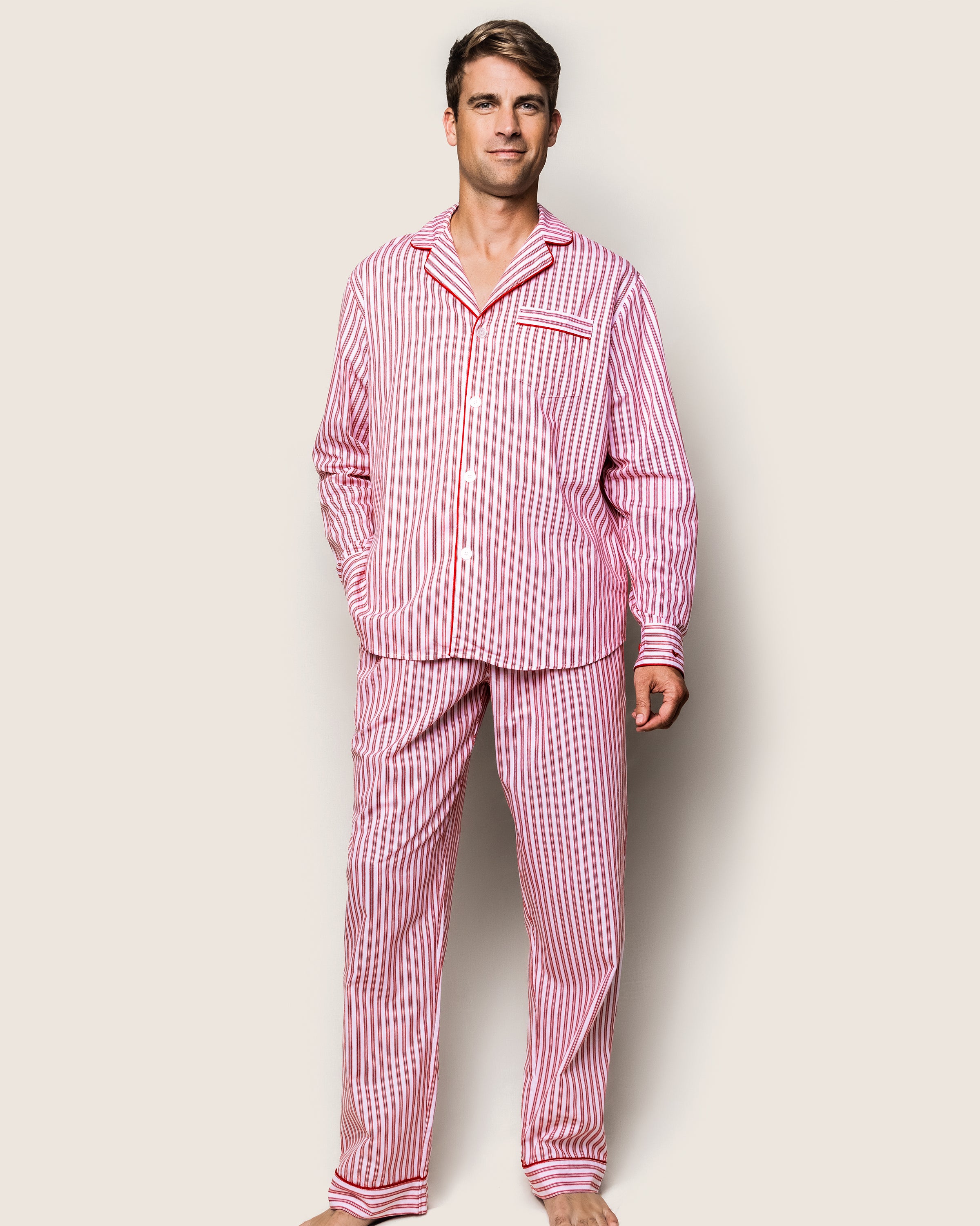A man stands barefoot against a plain background wearing the Petite Plume Mens Twill Pajama Set in Antique Red Ticking, featuring a red and white striped, moisture-wicking cotton twill fabric. The set includes a long-sleeved button-up top with a pocket, paired with matching pants.