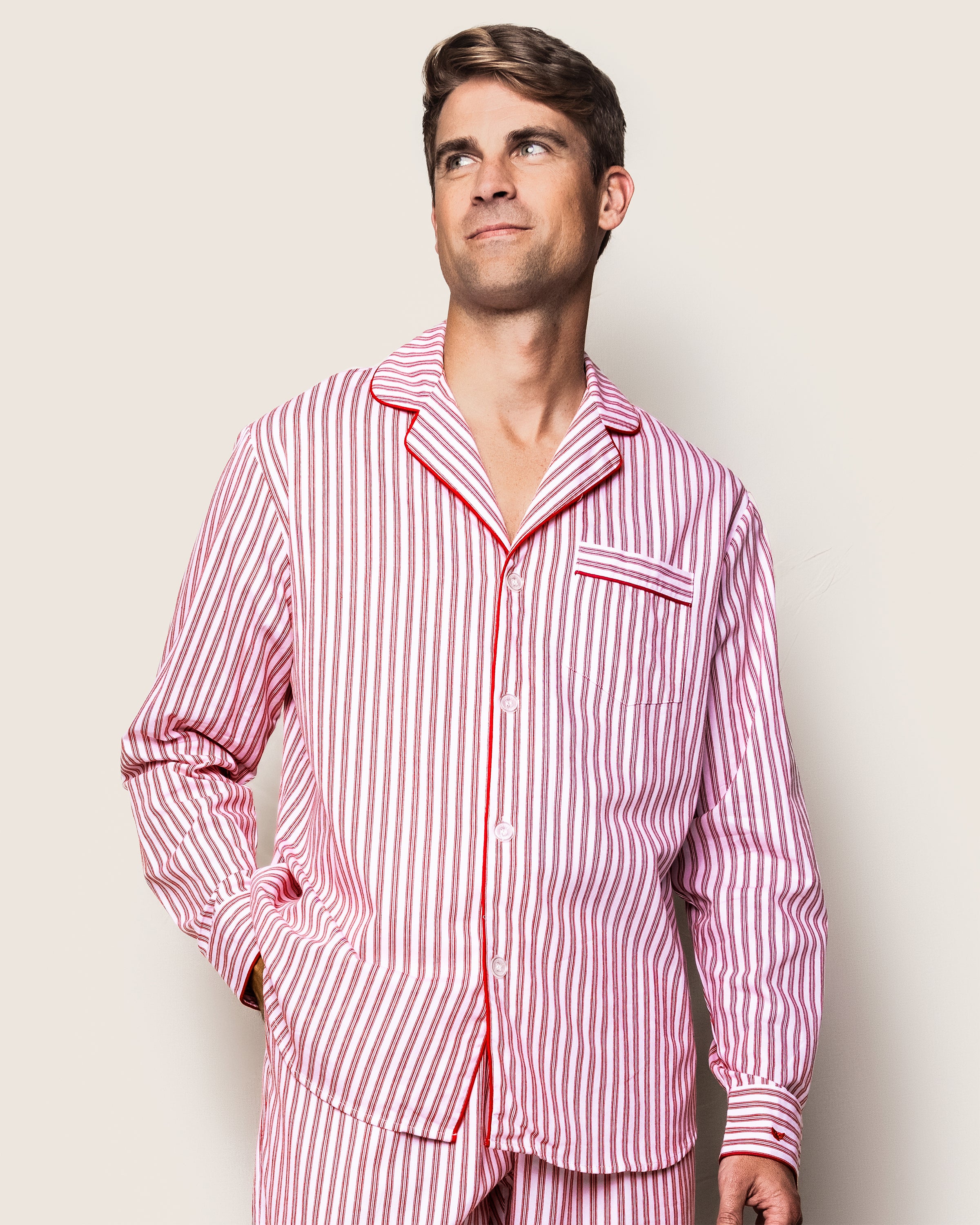 A man in Petite Plumes Mens Twill Pajama Set in Antique Red Ticking stands against a light backdrop. The 100% cotton twill top, featuring a pocket and buttons, offers a relaxed fit. He gazes upward with a slight smile.