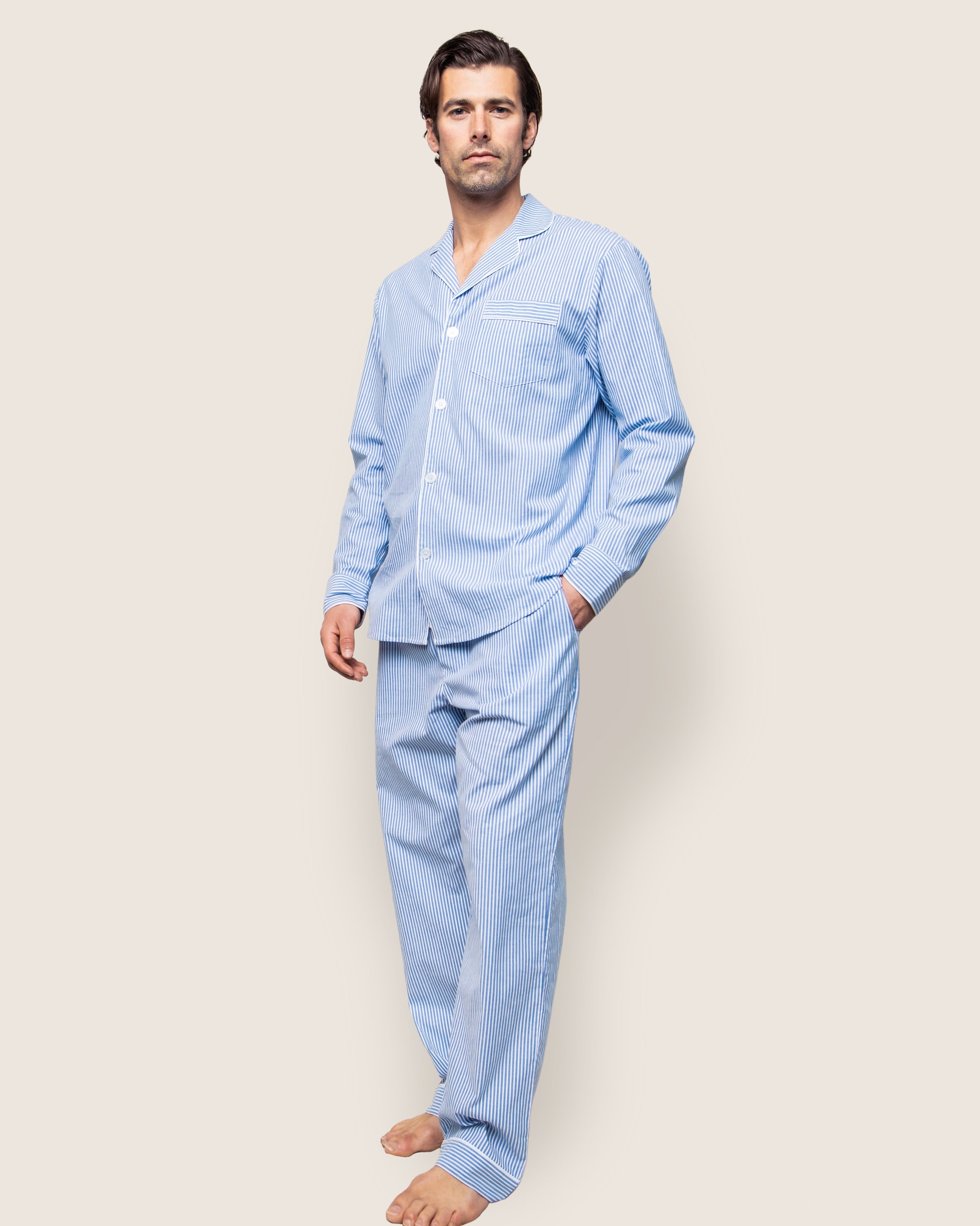 A man stands barefoot against a neutral background wearing Petite Plumes Mens Twill Pajama Set in French Blue Seersucker, which features a luxurious button-up top and long pants with chic light blue and white stripes, his left hand casually tucked in the pants pocket.
