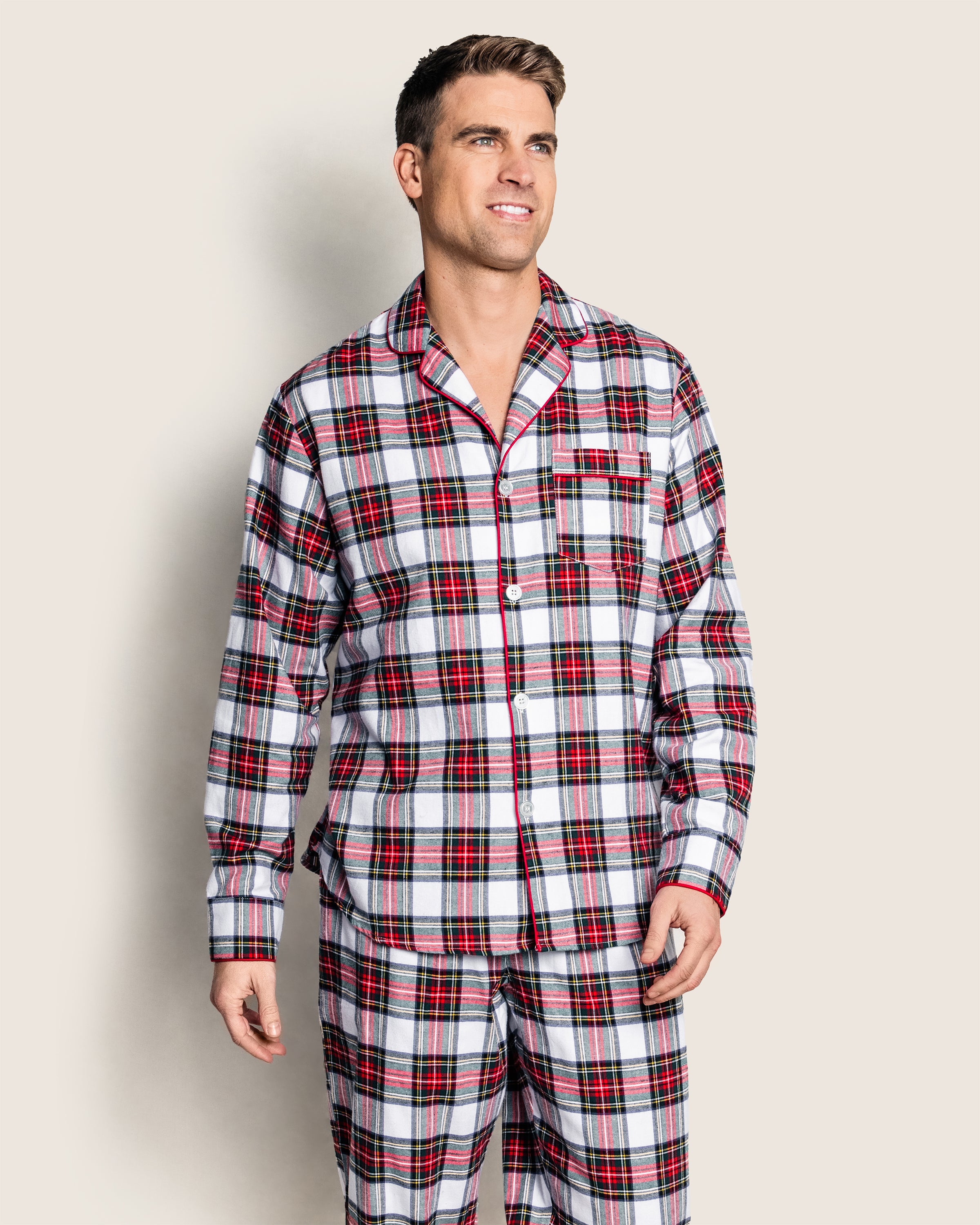 A person models Petite Plumes Mens Brushed Cotton Pajama Set in Balmoral Tartan, featuring a classic button-up shirt with a collar and pocket, paired with comfy pants, against a plain background.