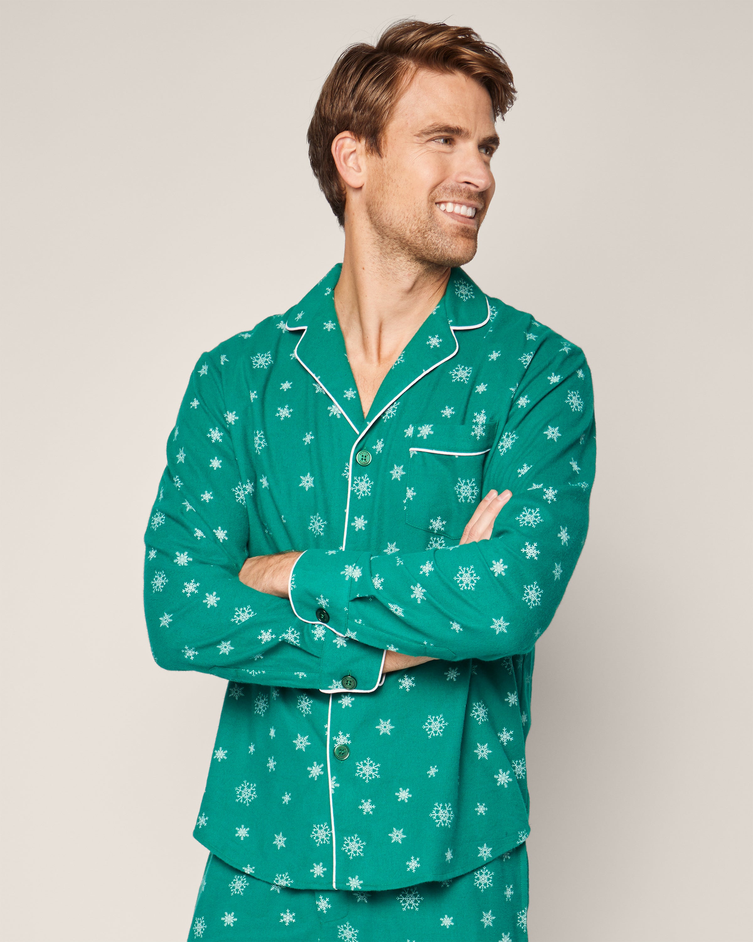 A smiling man wears the Mens Flannel Pajama Set in Emerald Wonderland by Petite Plume, featuring a classic emerald green design with white snowflakes. He stands with crossed arms, gazing sideways against a plain light-colored background.