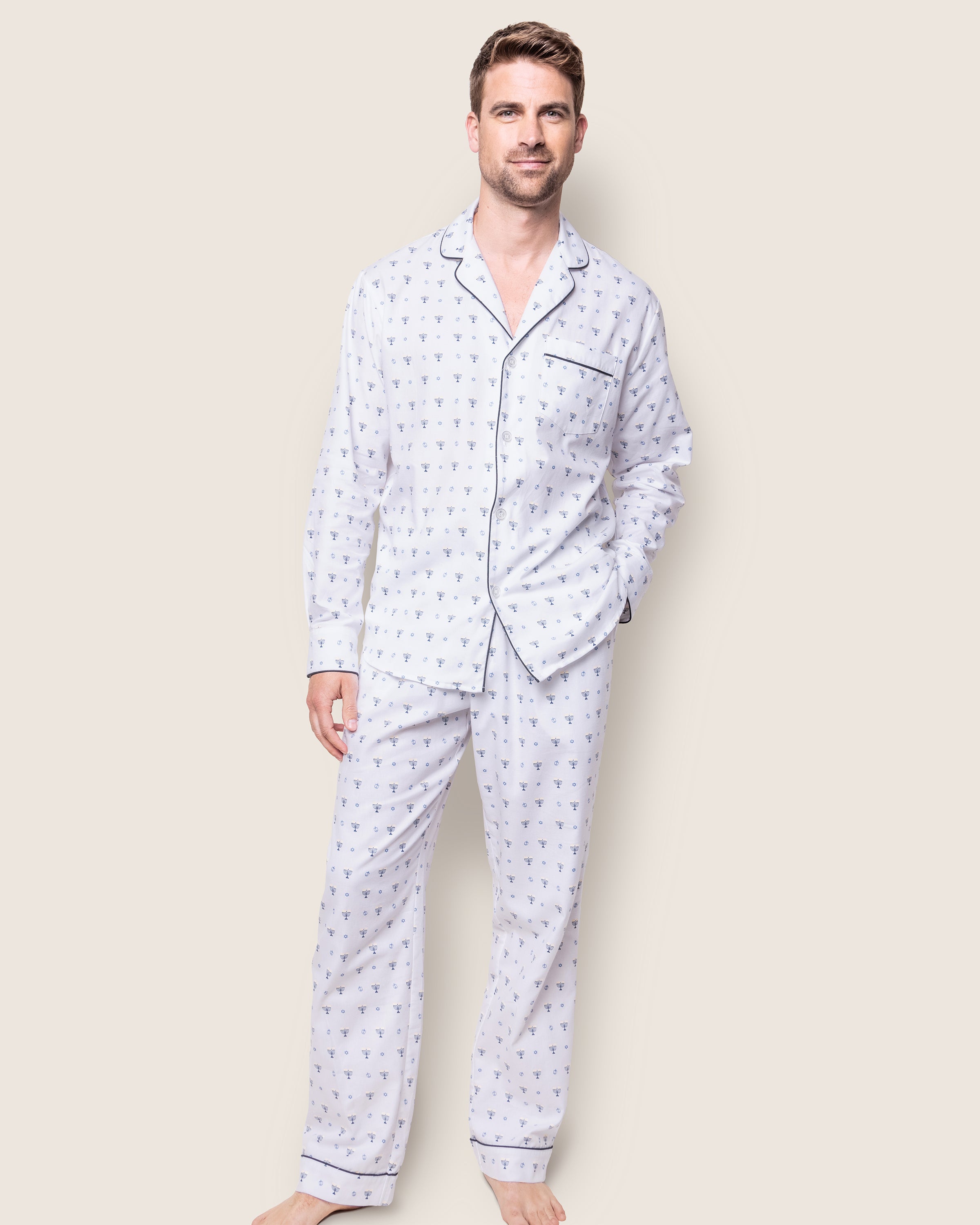 A man stands barefoot against a light background, wearing the Petite Plume Mens Twill Pajama Set in Happy Hanukkah. The white set features a subtle print with dark piping, and includes a long-sleeve button-up shirt with a pocket and matching pants.