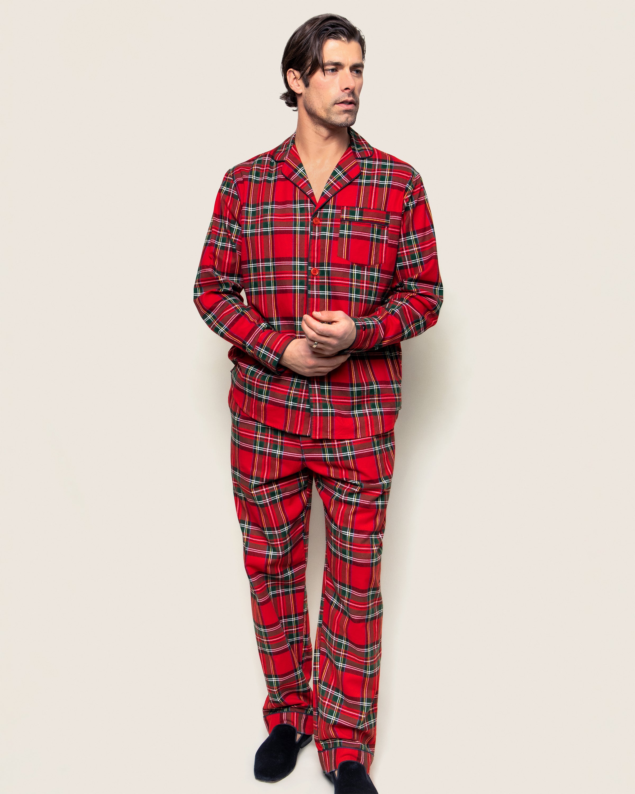 A person stands against a light background, wearing the Petite Plume Mens Brushed Cotton Pajama Set in Imperial Tartan with black slippers, looking to the side with hands together.