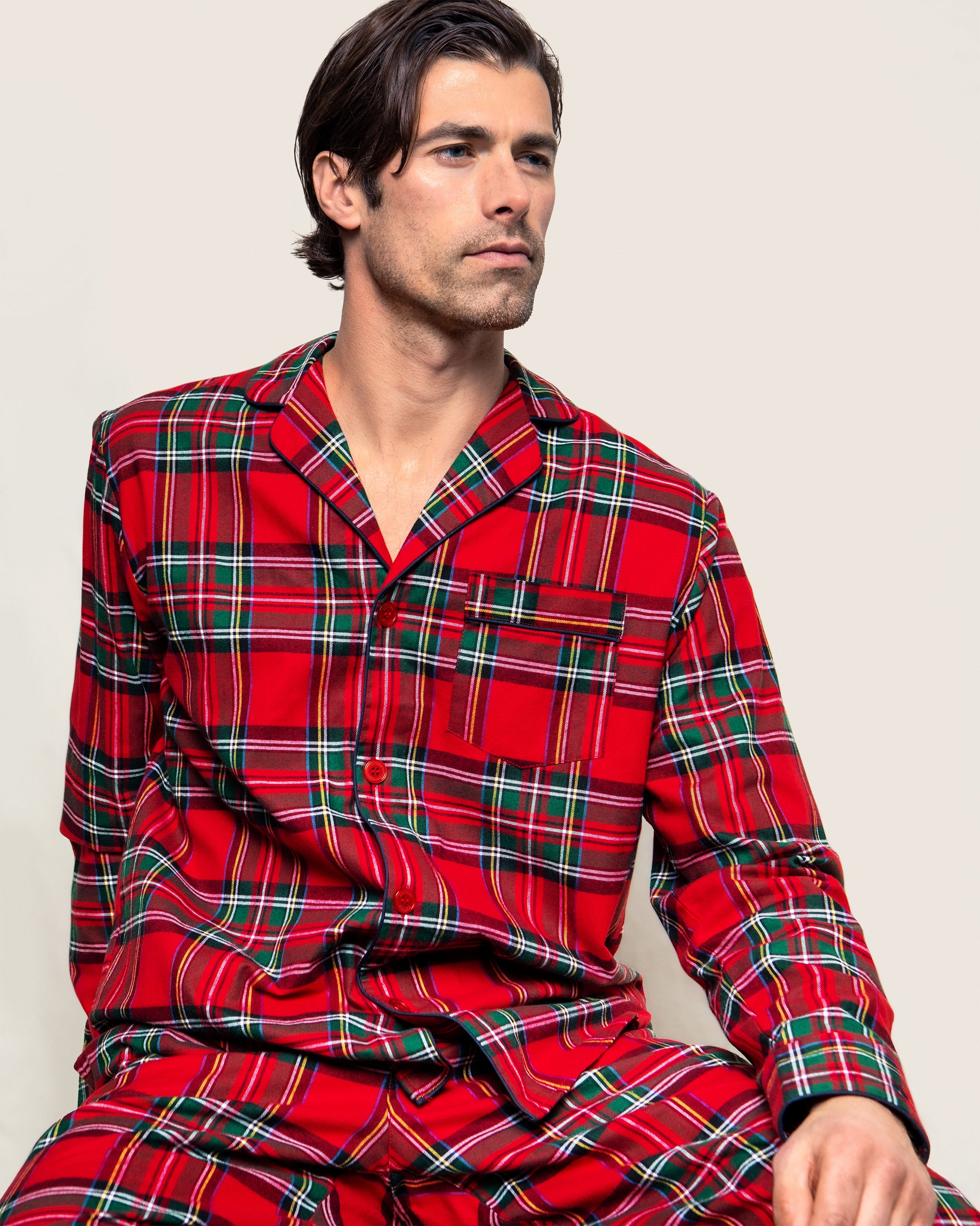 A person in a Petite Plume Mens Brushed Cotton Pajama Set, featuring an Imperial Tartan pattern, sits thoughtfully against a light background. The flannel sleepwear is button-up with a chest pocket.