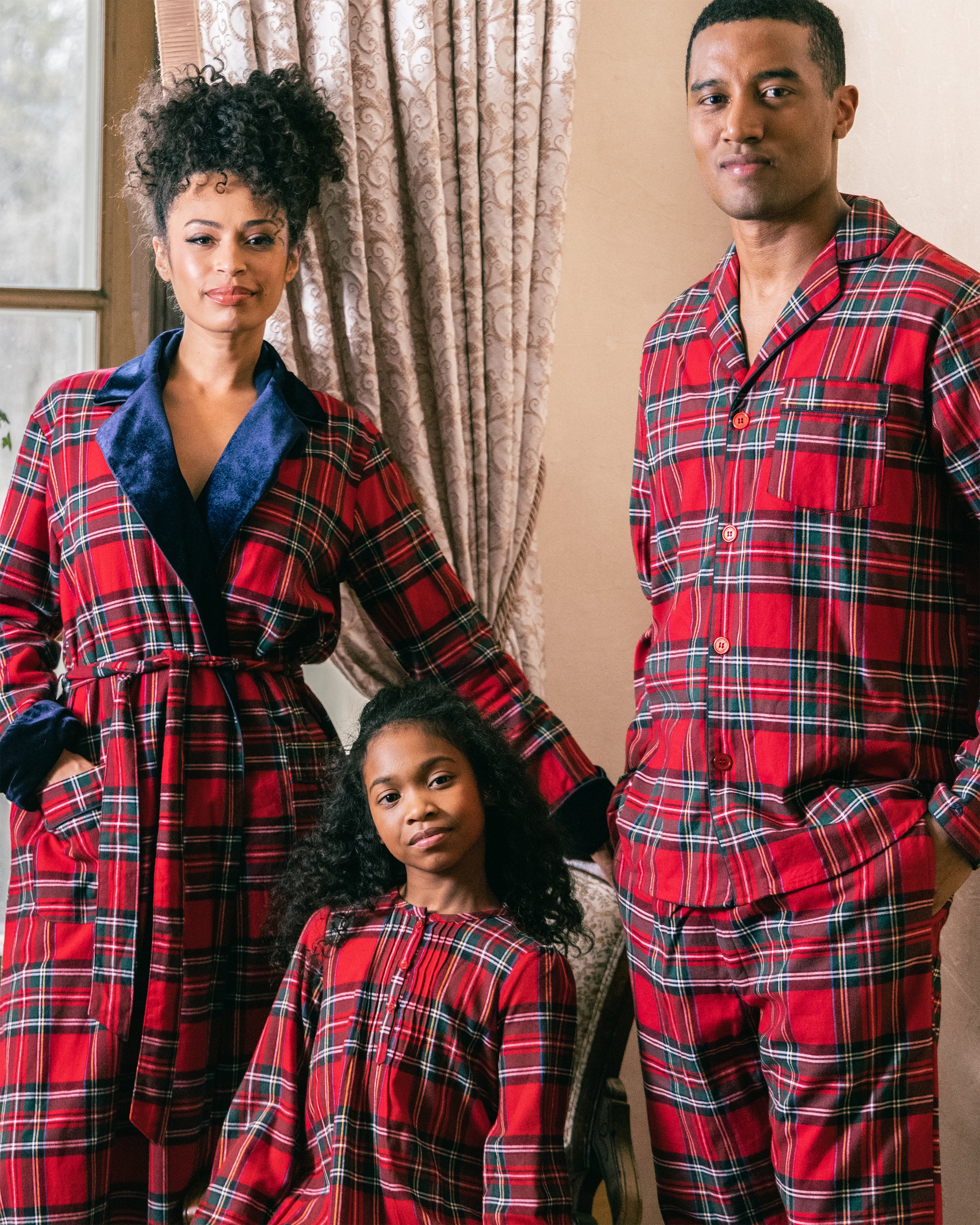 A family of three, all wearing Petite Plumes matching Imperial Tartan pajamas, stands indoors with a window and curtains behind them. The parents flank their daughter, looking content.