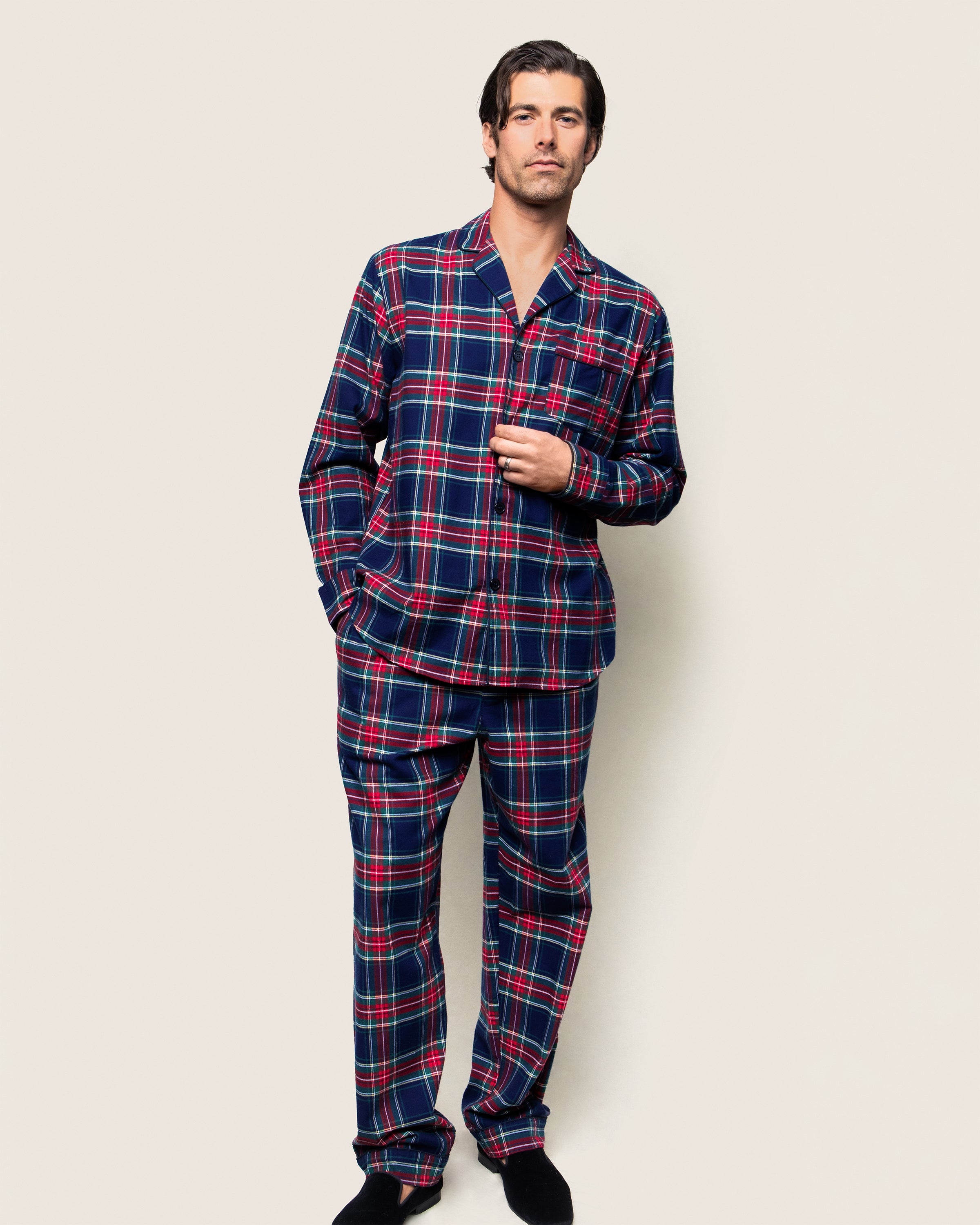 A man stands against a plain background wearing Petite Plumes Mens Brushed Cotton Pajama Set in Windsor Tartan, featuring lightweight flannel in red, blue, and green plaid. With one hand in his pocket and the other relaxed by his side, he completes the cozy look with black slippers.