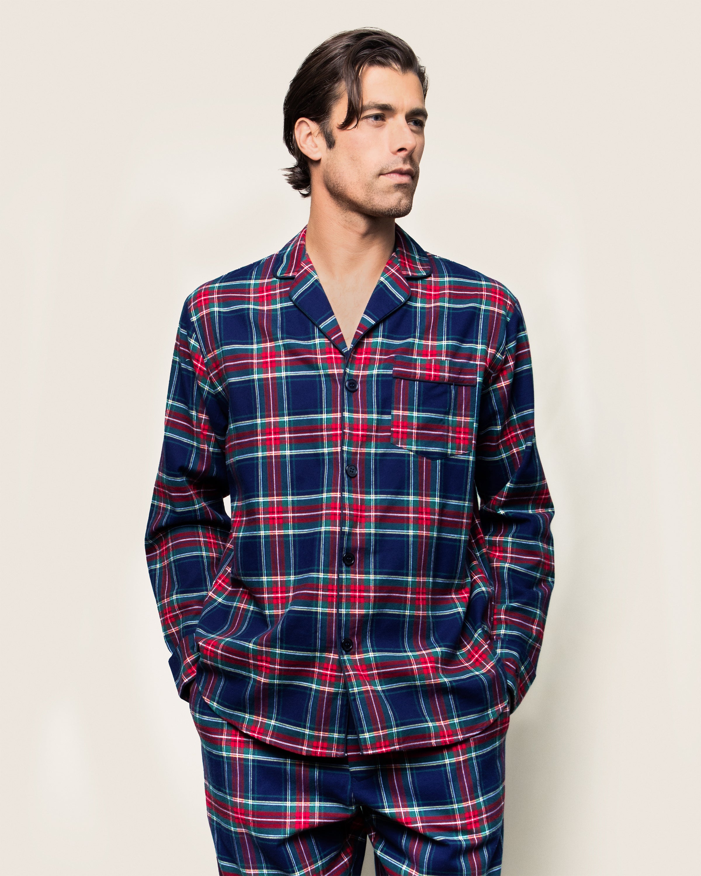 Wearing the Mens Brushed Cotton Pajama Set in Windsor Tartan by Petite Plume, someone stands against a plain light background. The high-quality cotton set includes a button-up top with a chest pocket. Gazing to the side, hands in pockets, they exude cozy elegance.