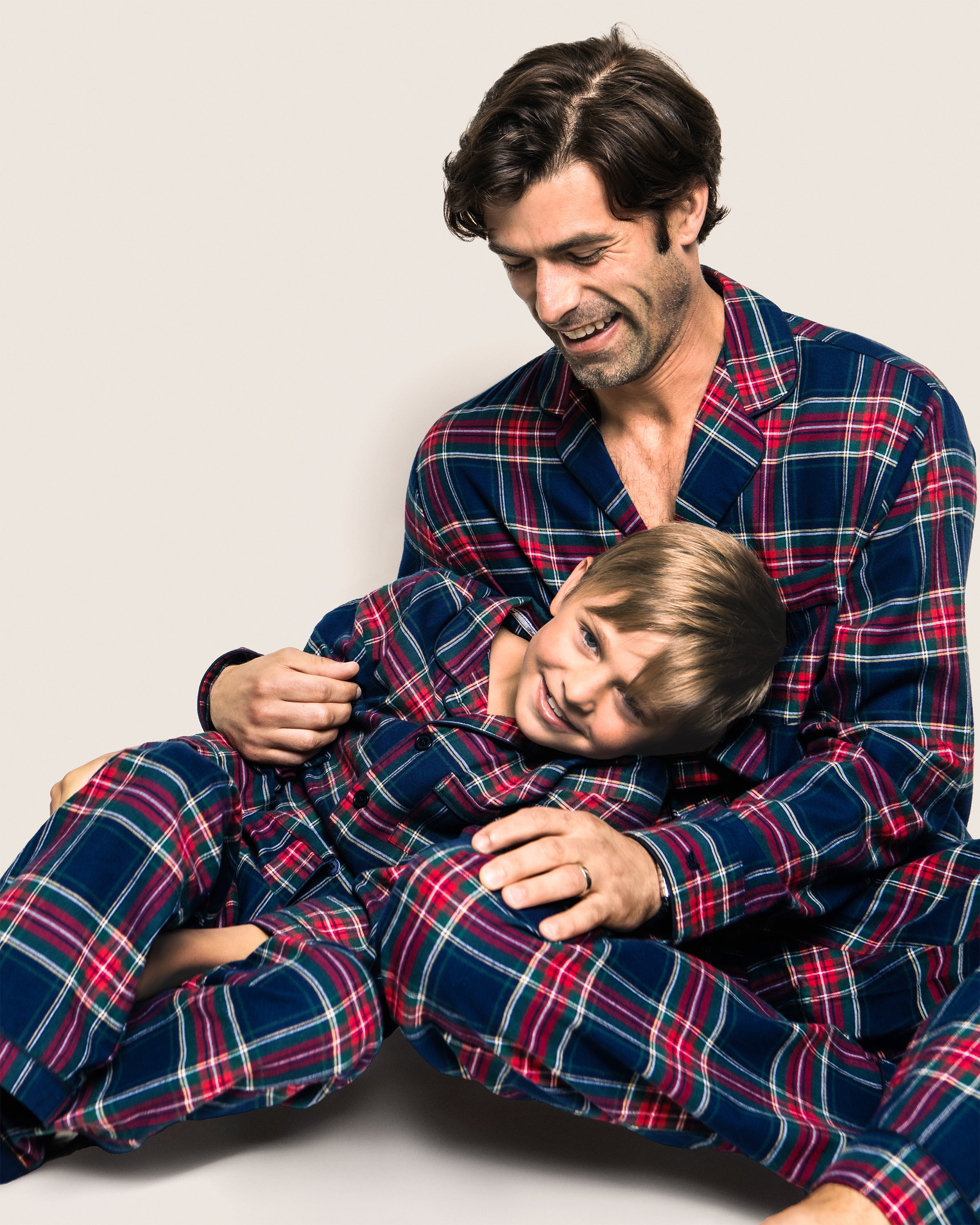 A man and child sit together in matching Petite Plume pajamas, made from brushed cotton. The man, wearing the Mens Windsor Tartan Set, smiles at the joyful child leaning against him. They are seated against a light background.