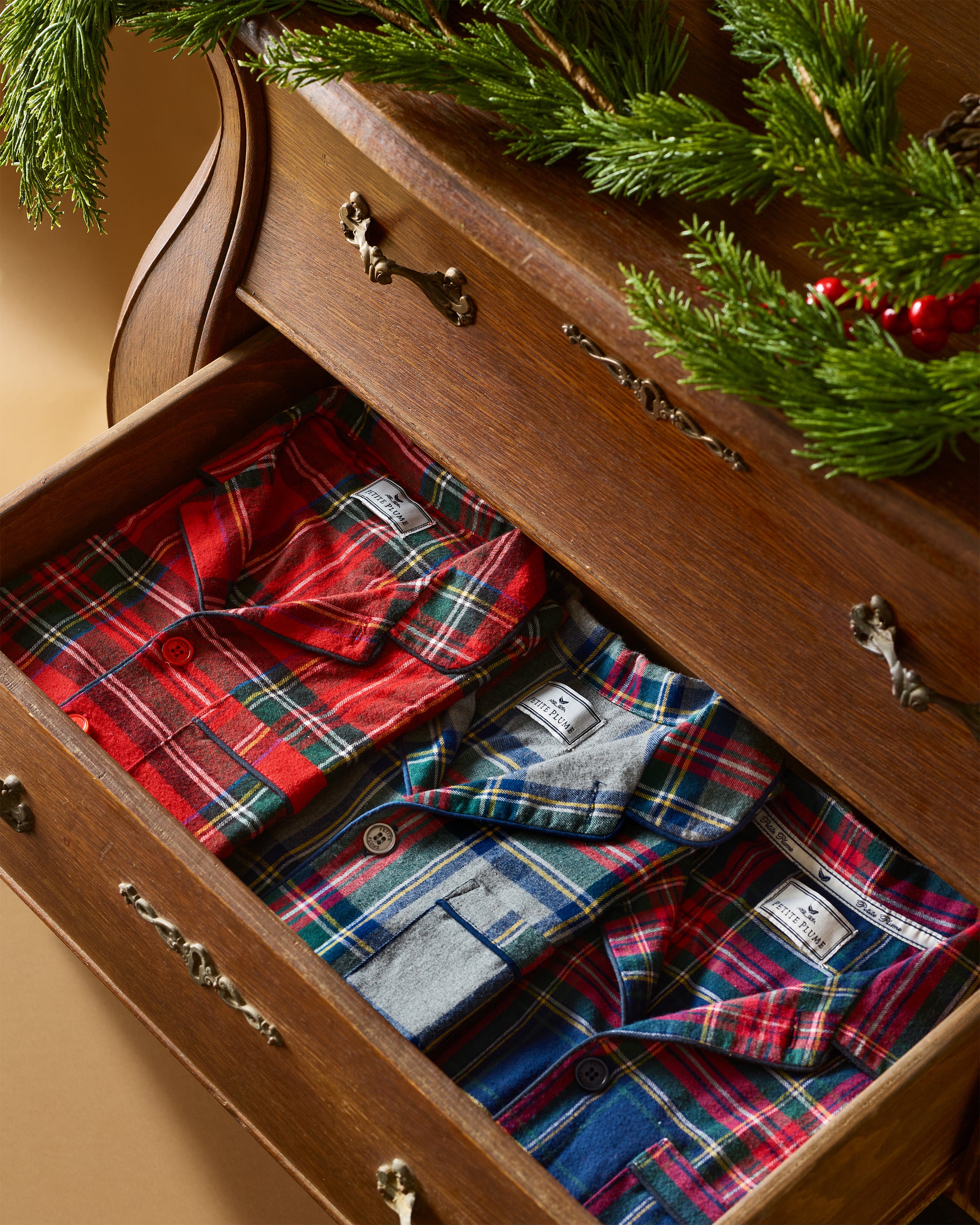 A wooden drawer displays three folded Petite Plume Mens Brushed Cotton Pajama Sets in Windsor Tartan, featuring plaid patterns in red, green, and blue. A sprig of green foliage with red berries decorates the drawers edge, creating a cozy, festive atmosphere.