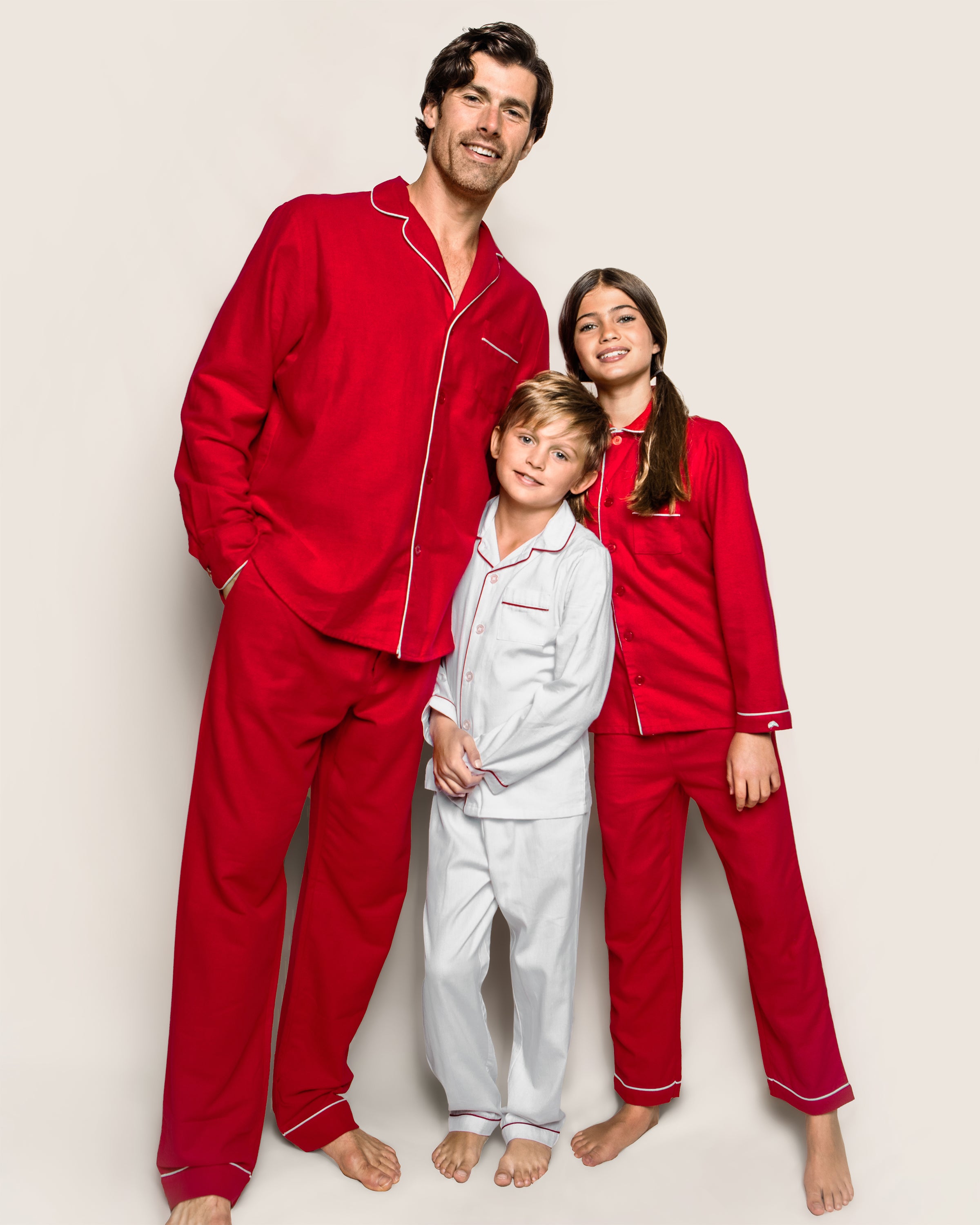 A man and two children smile together; the man and a girl wear matching Petite Plume Mens Flannel Pajama Sets in Red, while the boy sports crisp white pajamas. Barefoot against a plain background, they exude cozy luxury sleepwear vibes.