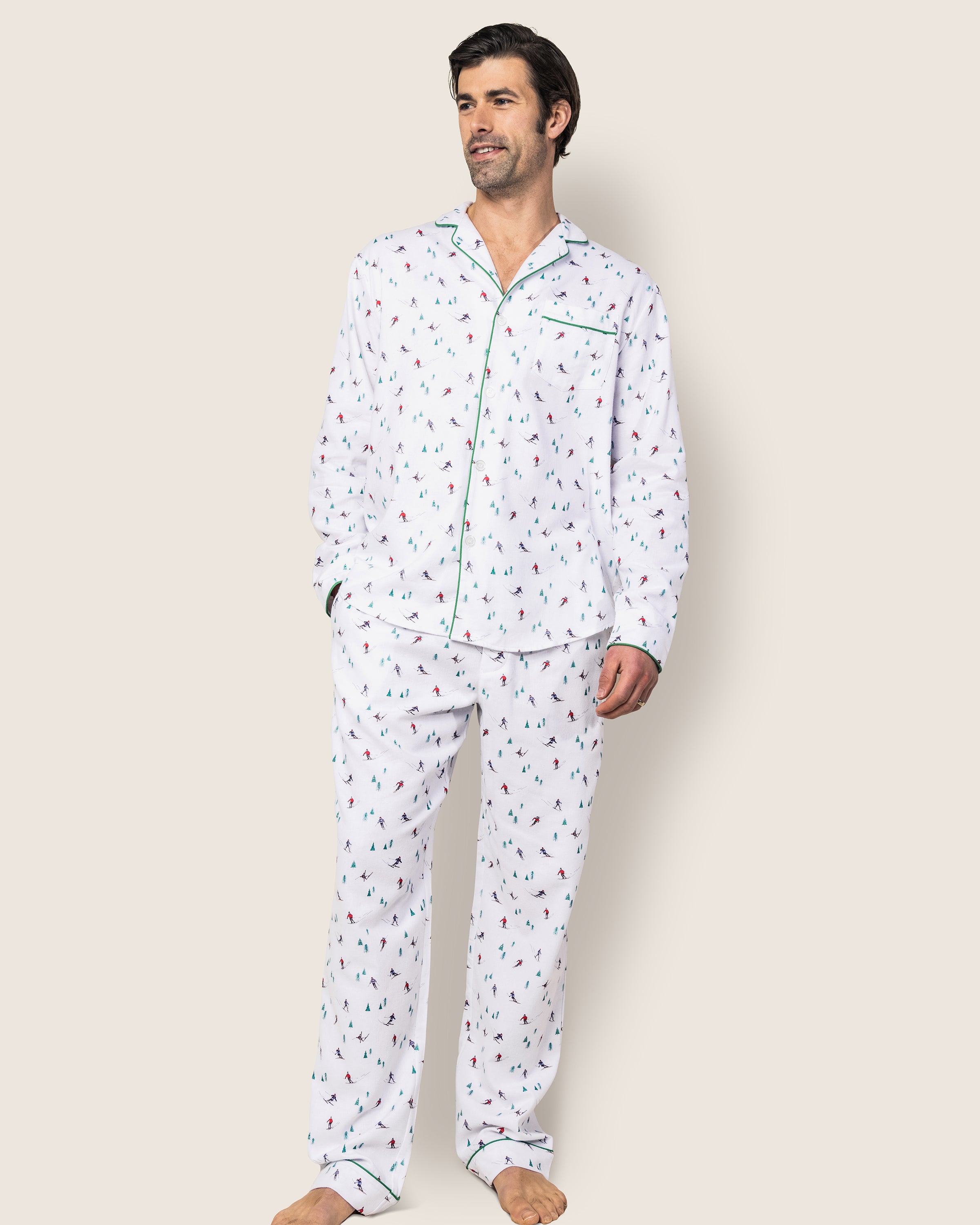 A man models the Mens Flannel Pajama Set in Après Ski by Petite Plume. Crafted from 100% cotton, it features a green-trimmed collar and cuffs. Against a plain background, he stands barefoot with one hand in his pocket, gazing slightly to the side.