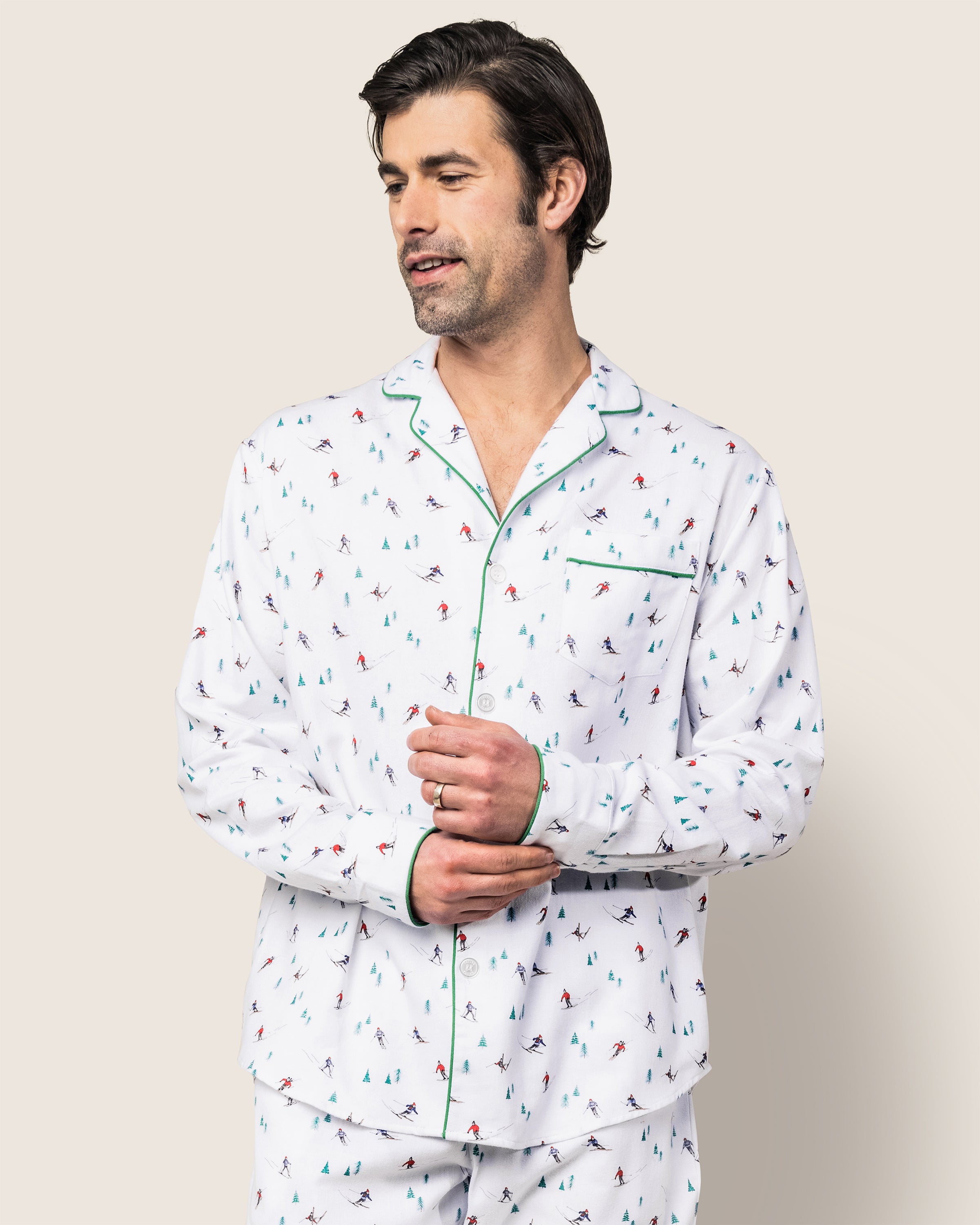 A man models the Mens Flannel Pajama Set in Après Ski by Petite Plume. The classic white sleepwear showcases colorful skier prints and features green piping along the collar and pocket, adding a timeless touch to this luxury set against a plain background.