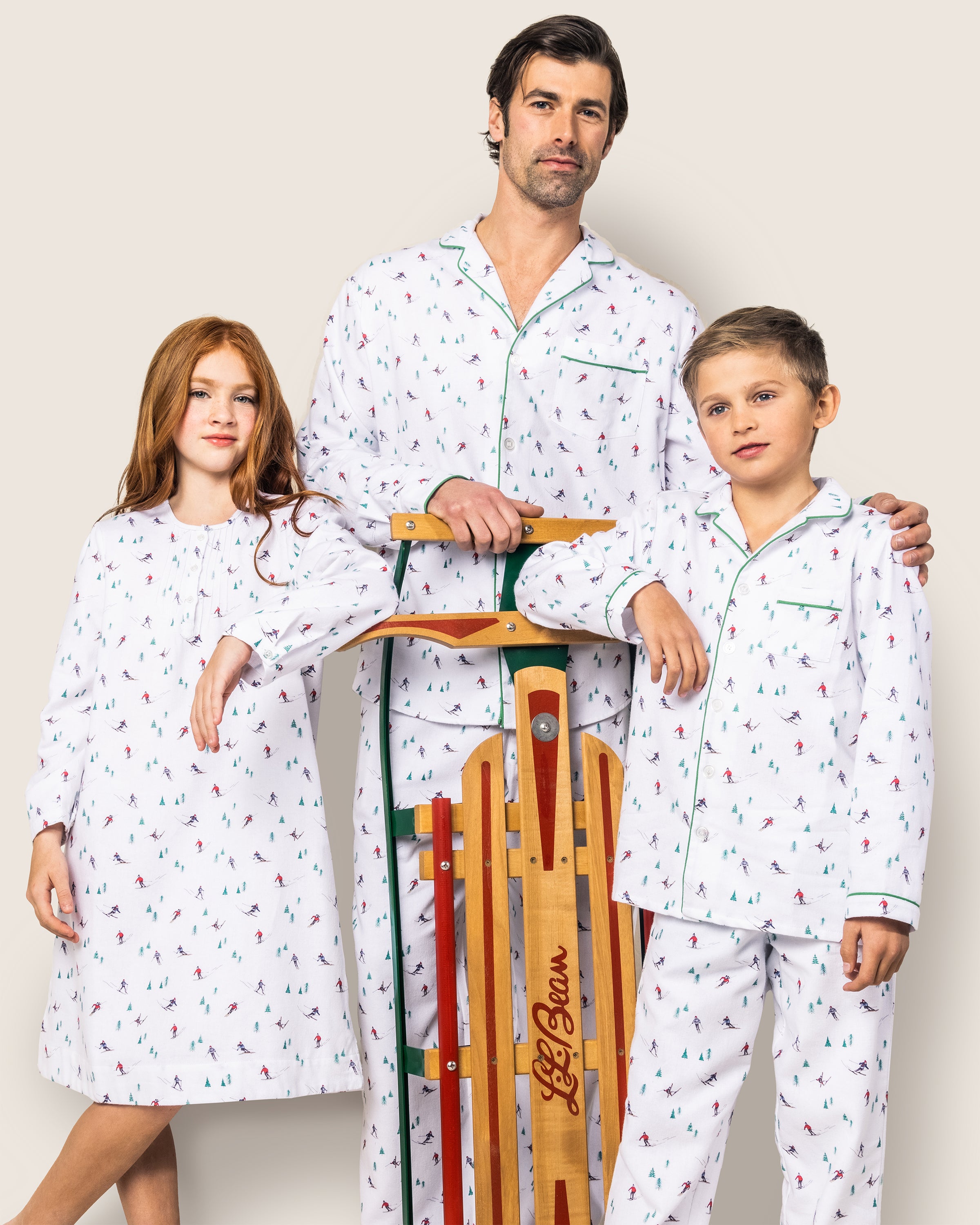 A man and two kids pose with a wooden sled, all wearing Petite Plumes Mens Flannel Pajama Set in Après Ski. The girls red hair contrasts with their festive 100% cotton pajamas, while the boy shares brown hair similar to the mans short dark hair and facial hair.