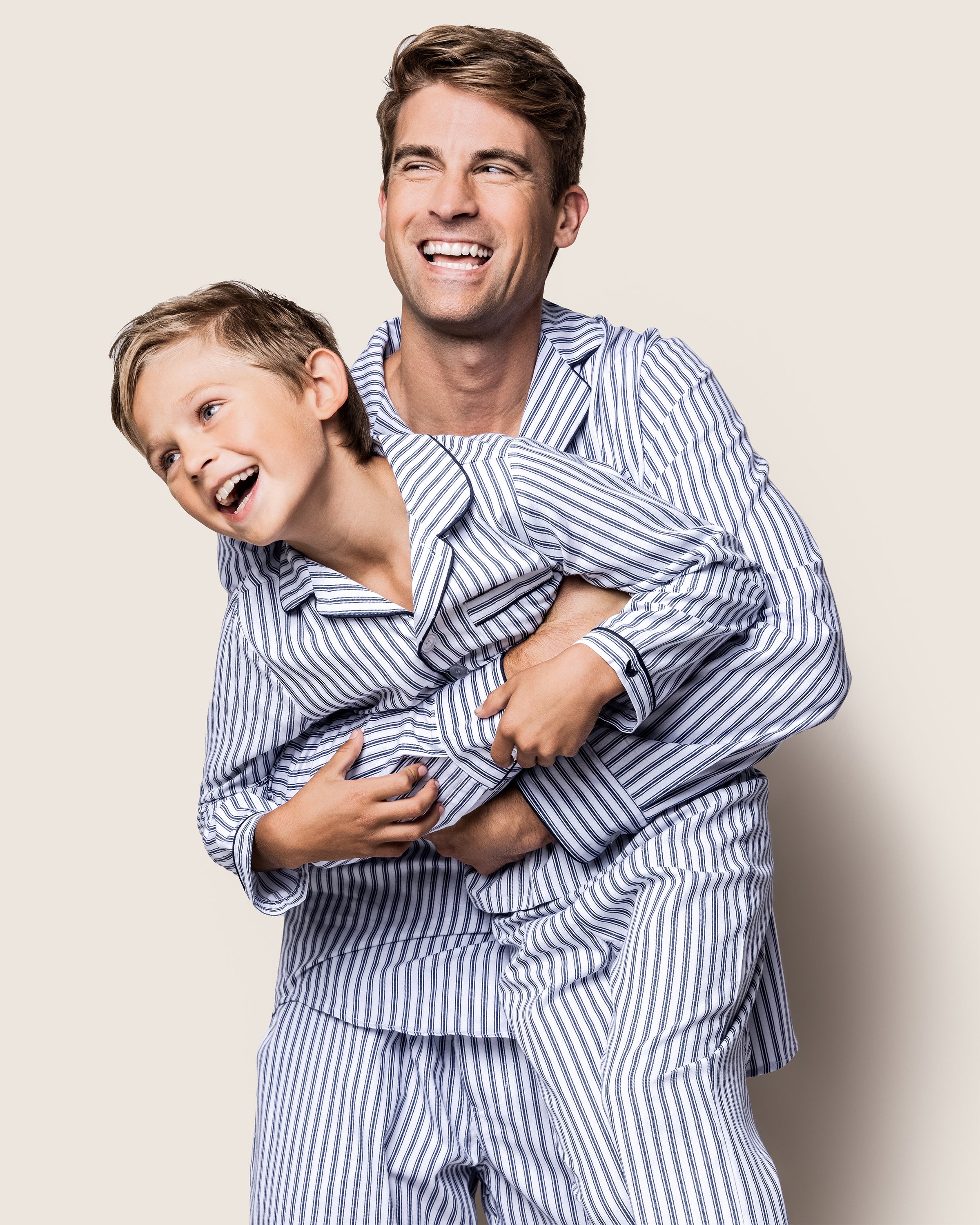 A man and child, wearing the Petite Plume Mens Twill Pajama Set in Navy French Ticking, are sharing laughs and hugs. With the mans arms around the childs waist, these 100% cotton pajamas enhance their joy against a simple light backdrop.