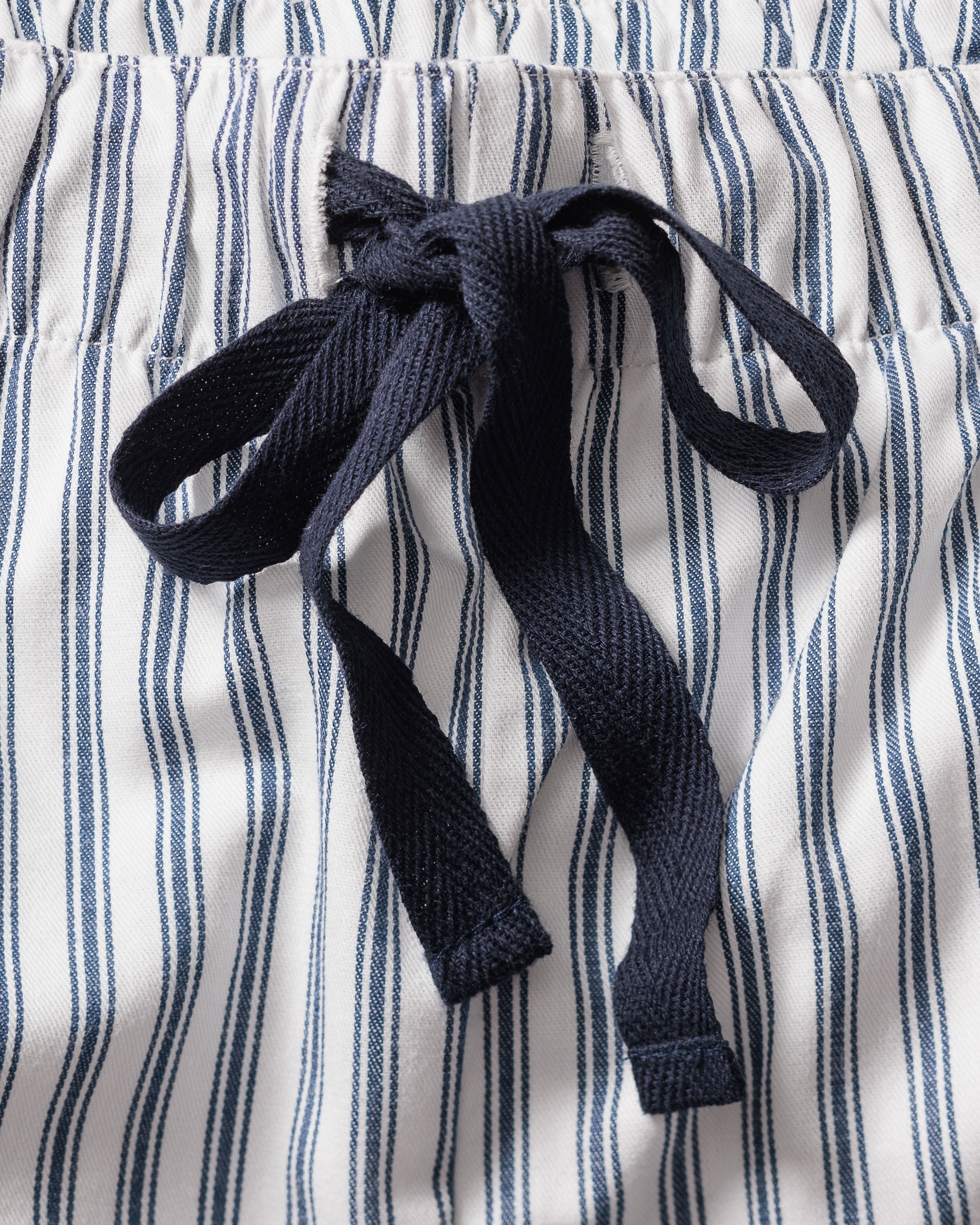 Close-up of the Mens Twill Pajama Set in Navy French Ticking by Petite Plume, featuring blue and white vertically striped shorts with an elastic waistband and a dark blue drawstring bow, highlighting their cozy 100% cotton feel.