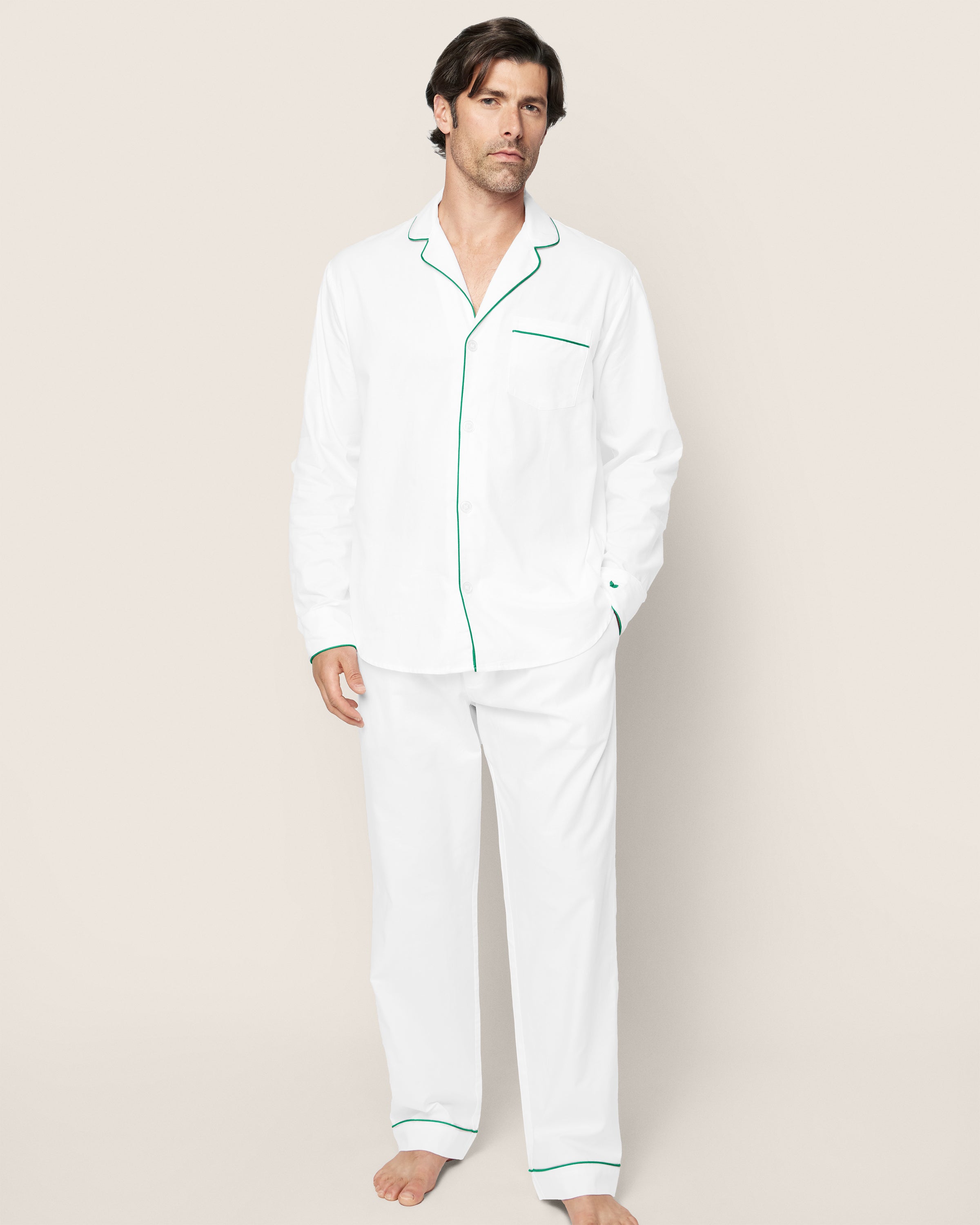 A person is barefoot against a plain background, wearing the Petite Plume Mens Twill Pajama Set in white with green piping. Crafted from high-quality cotton, it features a button-up long-sleeve top and matching pants.