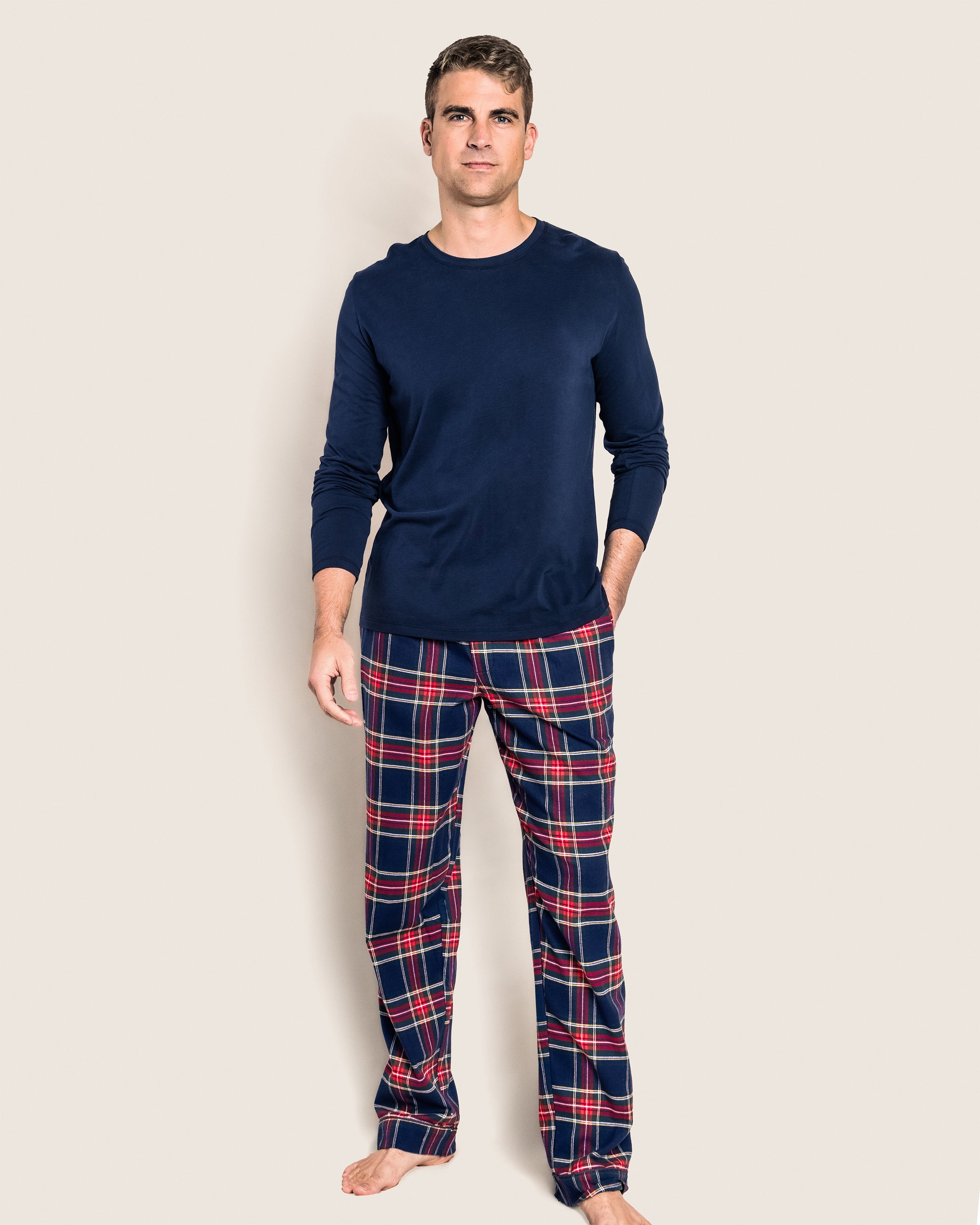 A person stands against a plain background wearing a navy long-sleeve shirt and Petite Plumes Mens Brushed Cotton Pants in Windsor Tartan, showcasing red, blue, and white plaid. This cozy sleepwear set is made from 100% cotton. They are barefoot and facing the camera.