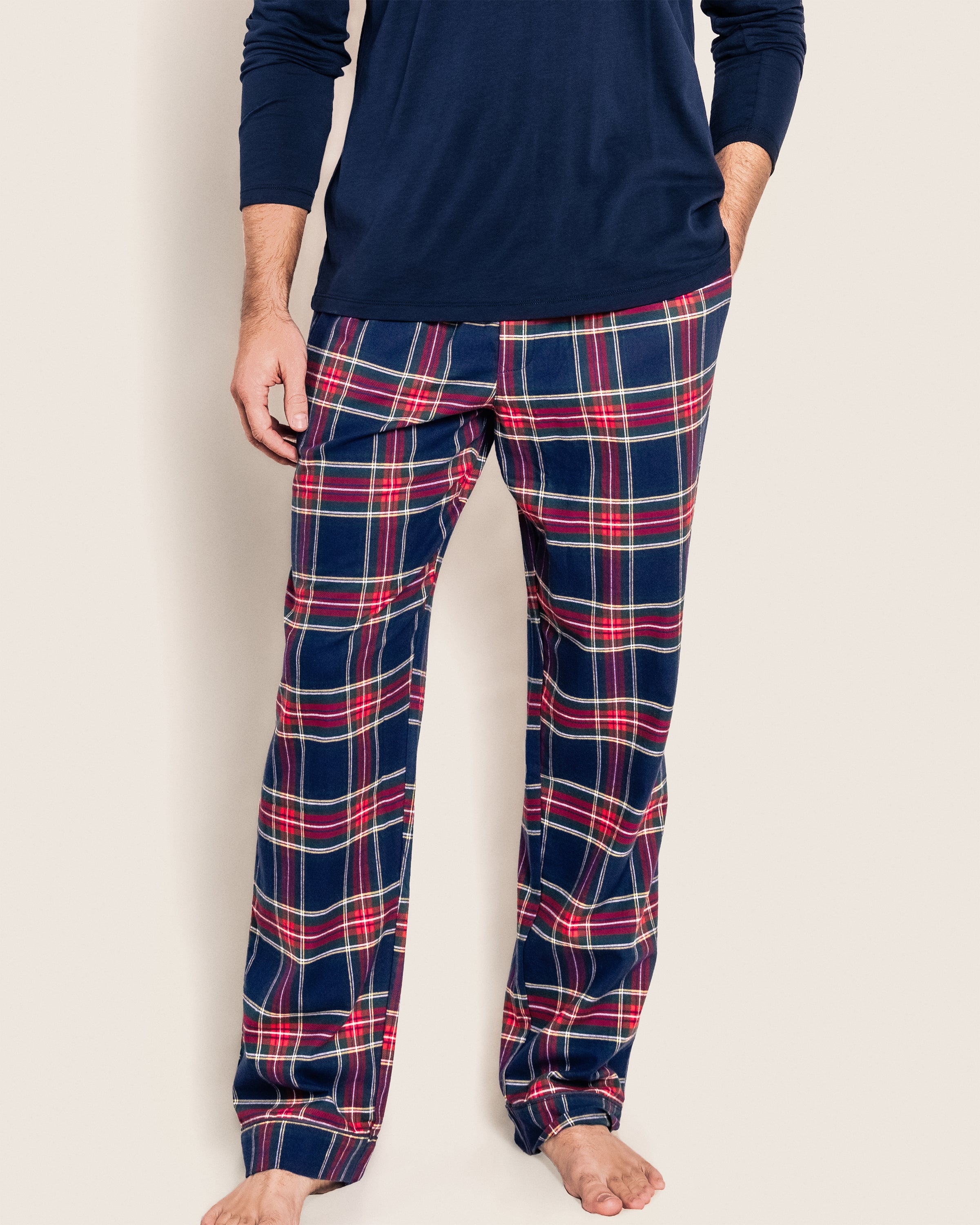 Dressed in Petite Plumes Mens Brushed Cotton Pants in Windsor Tartan, featuring a plaid design of red, white, and navy blue lines, paired with a long-sleeved navy shirt, a person stands barefoot against an off-white background. The image captures from shoulders down.