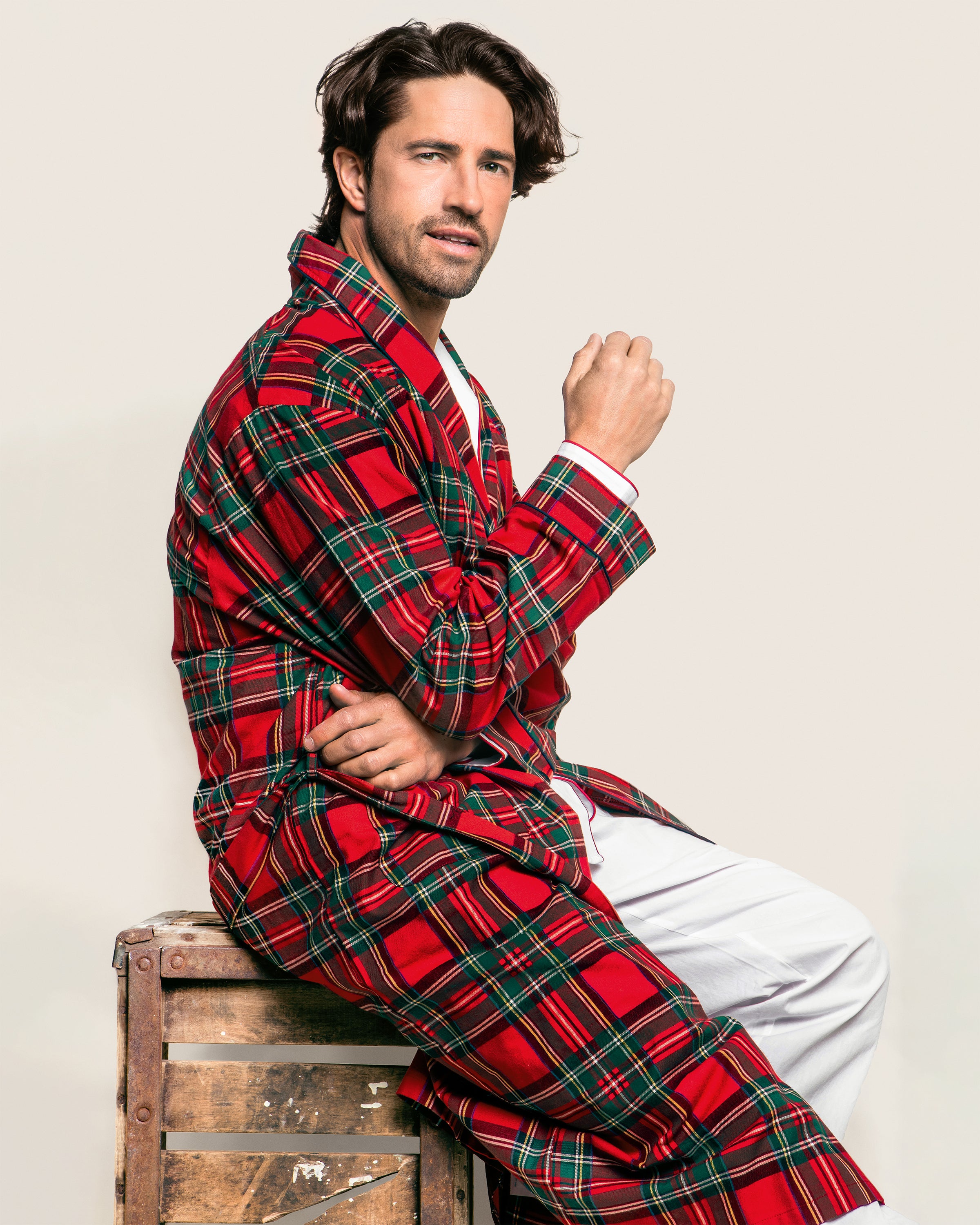 A bearded man with dark hair, wearing a Petite Plume Mens Brushed Cotton Robe in Imperial Tartan over white pants, sits on a wooden crate against a light beige background. He is slightly angled to the side, with one hand near his chin.