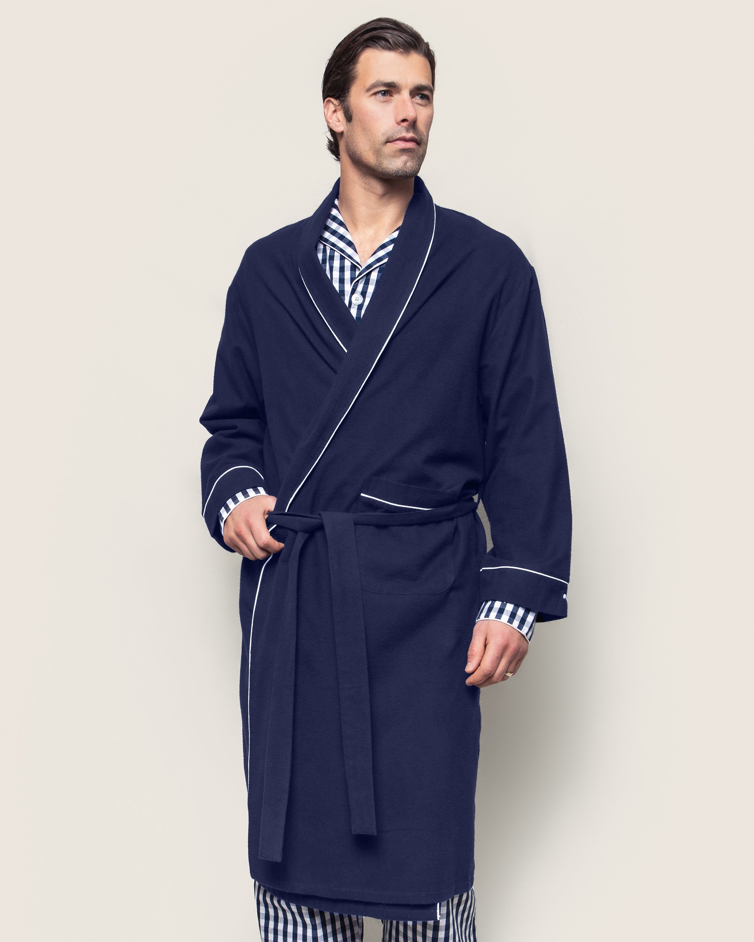 A man wears Petite Plumes Mens Flannel Robe in Navy with White Piping, featuring moisture-wicking fabric and a tie belt over striped pajamas. He stands against a plain background, gazing to the side with a calm expression.