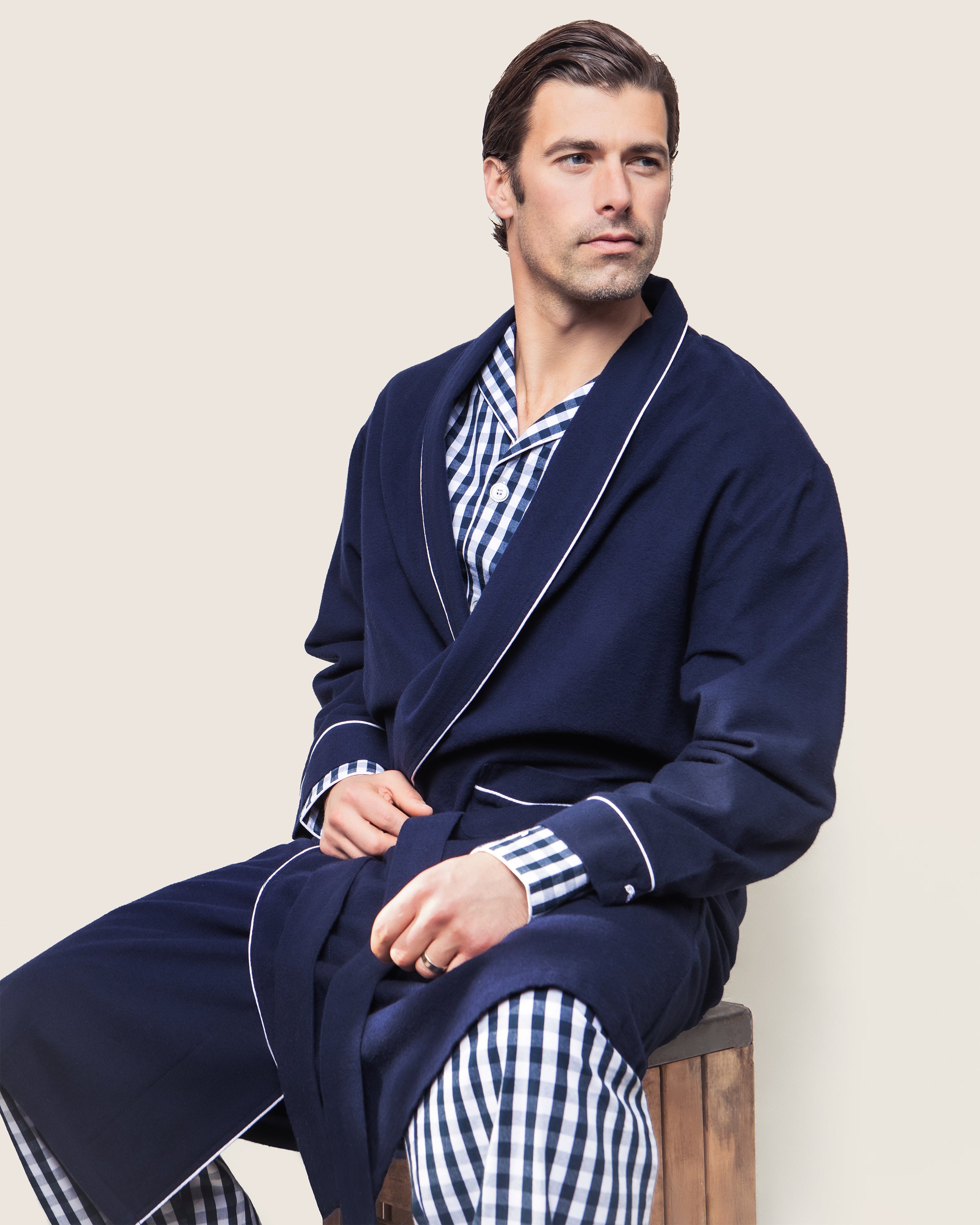 A person wearing Petite Plumes Mens Flannel Robe in Navy with White Piping over blue and white checkered flannel pajamas sits on a wooden stool against a plain background, looking to the side.