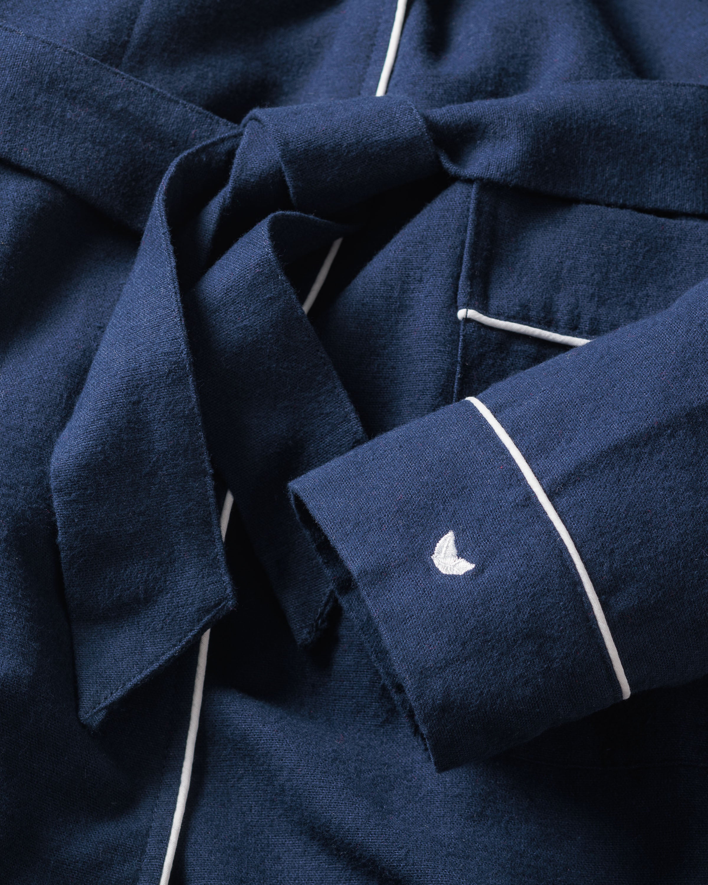 Close-up of Petite Plumes Mens Flannel Robe in Navy with white piping. Features a tied belt and a small white crescent logo on the sleeve. The moisture-wicking fabric looks soft and textured.