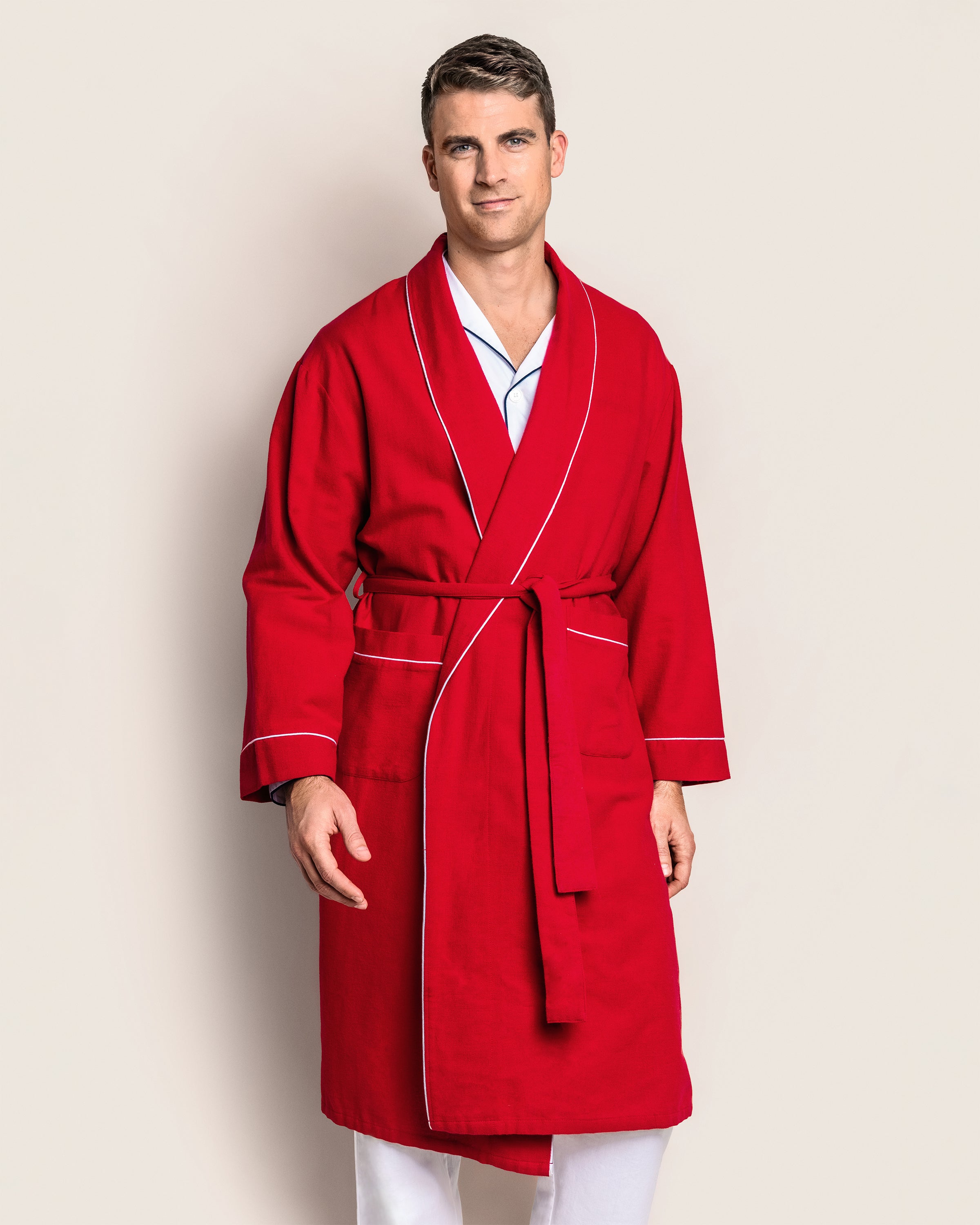 A man wears a Petite Plume Mens Flannel Robe in Red with White Piping, tied at the waist over his white pajamas. Crafted from soft cotton, he stands against a neutral background, gazing at the camera.