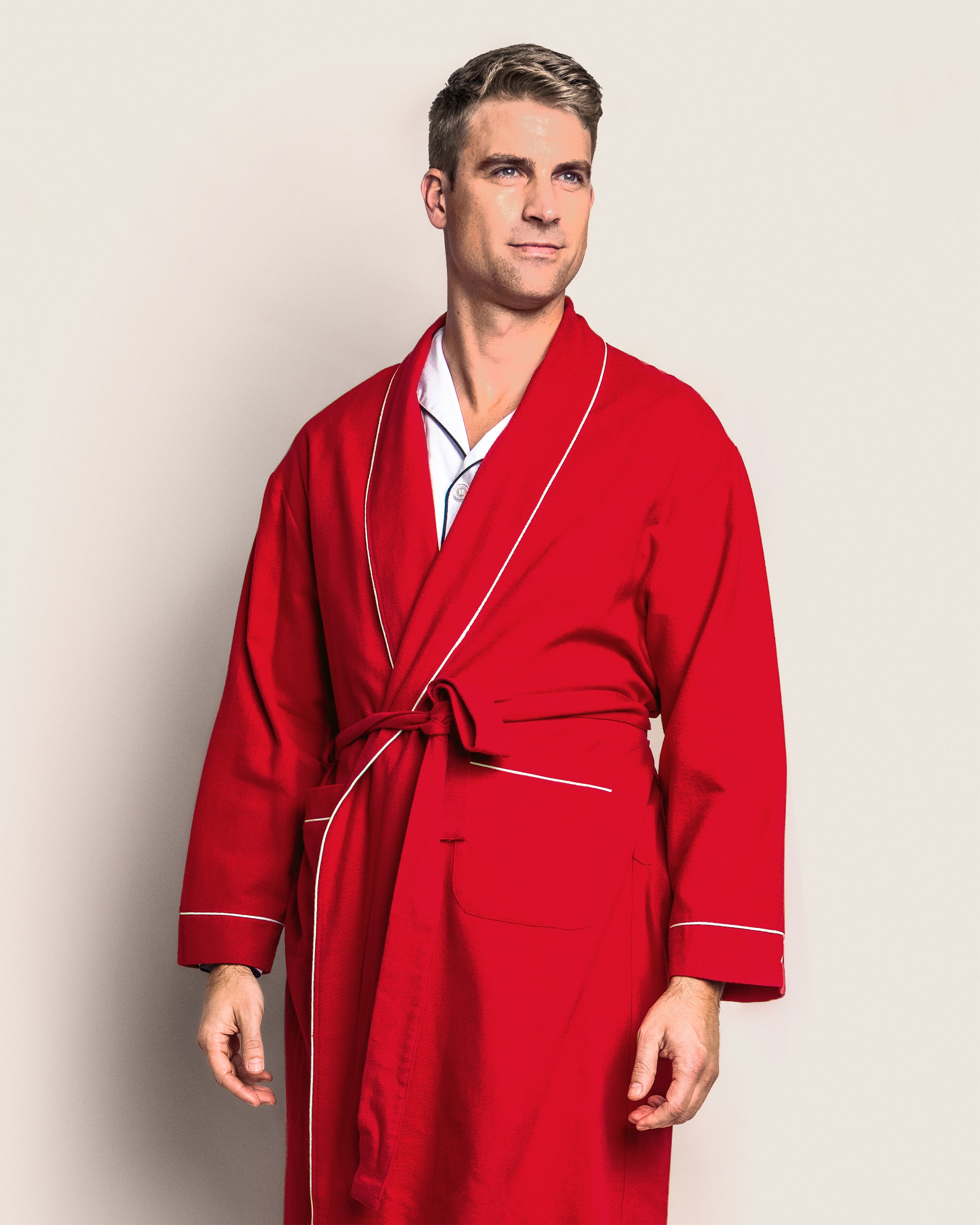 A person in a luxurious Mens Flannel Robe in Red with White Piping by Petite Plume stands against a light background, looking slightly to the side with one hand by their side and the other slightly bent.