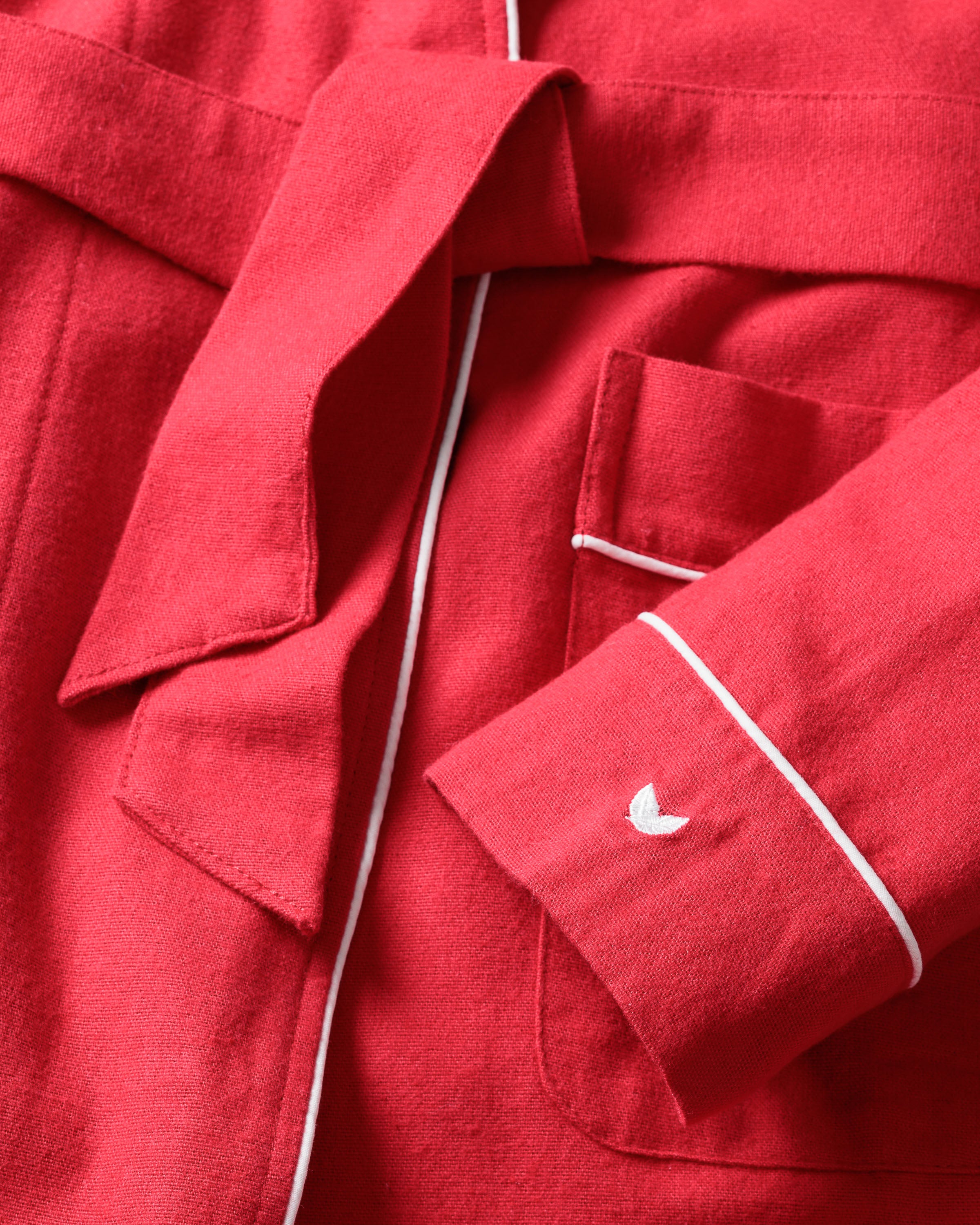 The Petite Plume Mens Flannel Robe in Red, perfect for luxury sleepwear, features white piping, a belt, large front pockets, and a small embroidered white crescent on the sleeve. The cotton fabric has a soft and textured appearance.