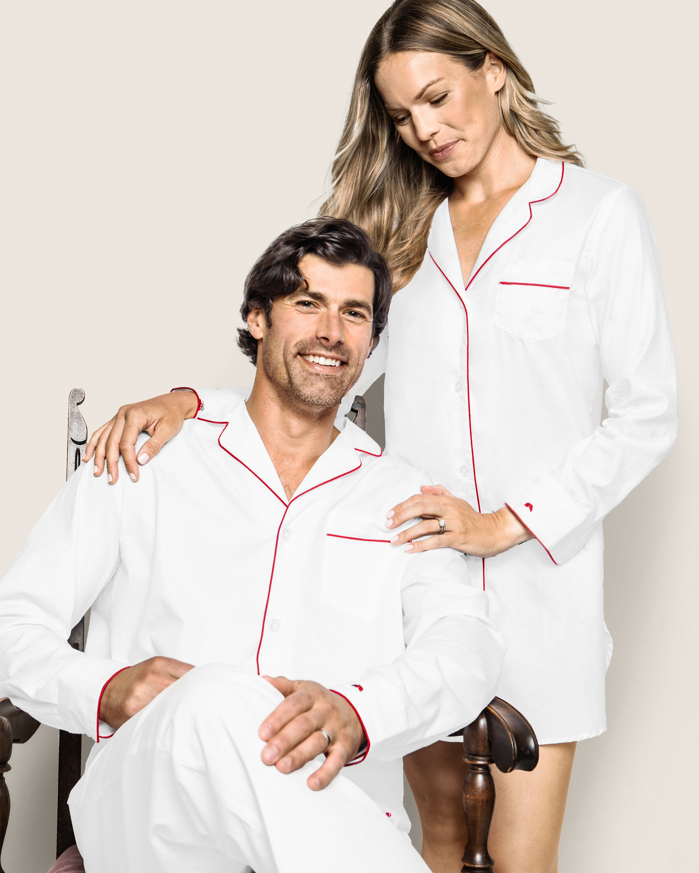 A man sits in a wooden chair, wearing white pajamas with red piping, smiling at the camera. A woman stands beside him, clad in a Petite Plume Womens Twill Nightshirt in White with Red Piping, smiling gently. Their arms rest casually on each other, capturing a moment of relaxed comfort.