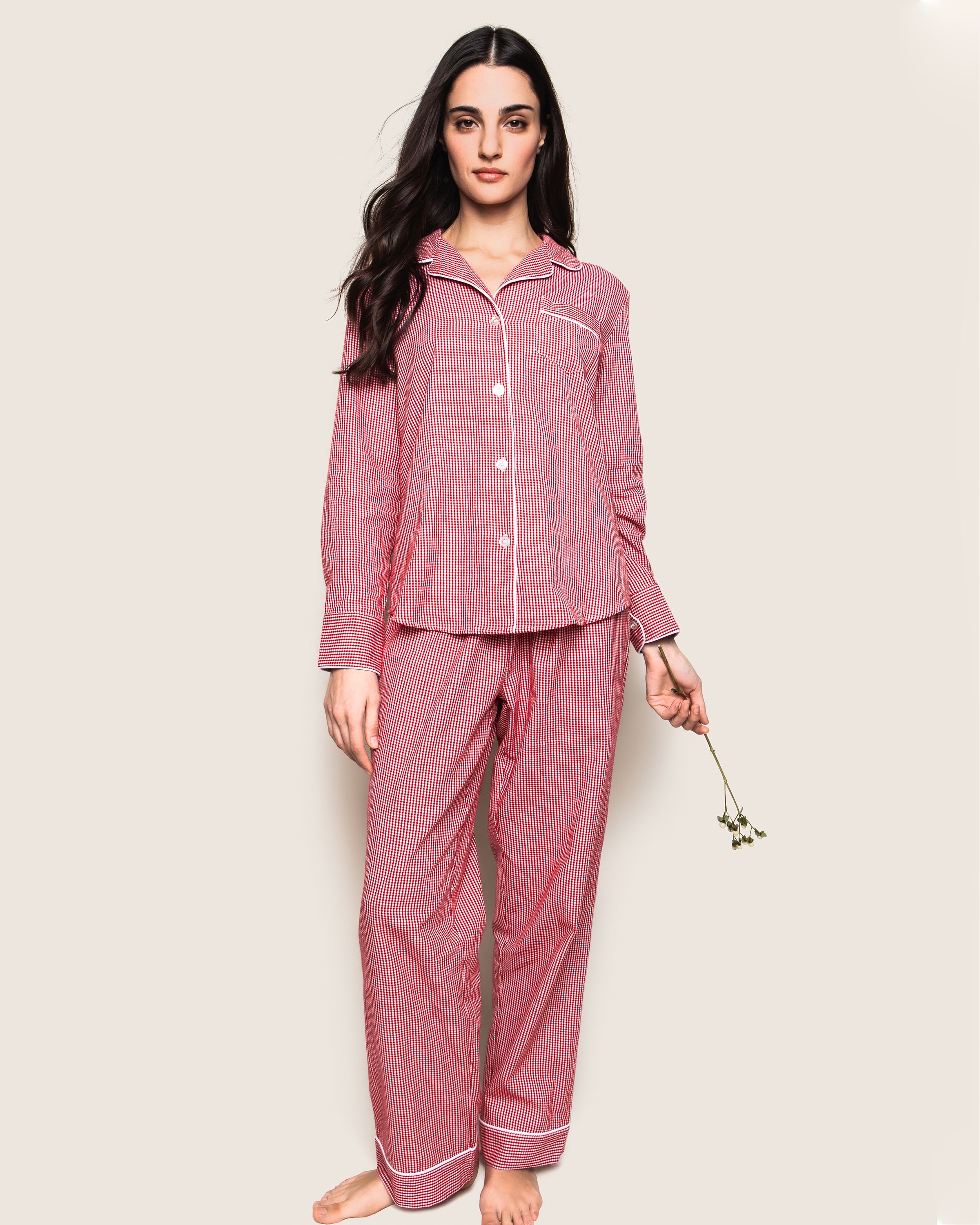 A woman stands barefoot against a plain background, wearing the Womens Flannel Pajama Set in Red Mini Gingham by Petite Plume. She holds small flowers in her right hand, her long dark hair cascading over her shoulders—combining comfort and elegance unique to luxury sleepwear.