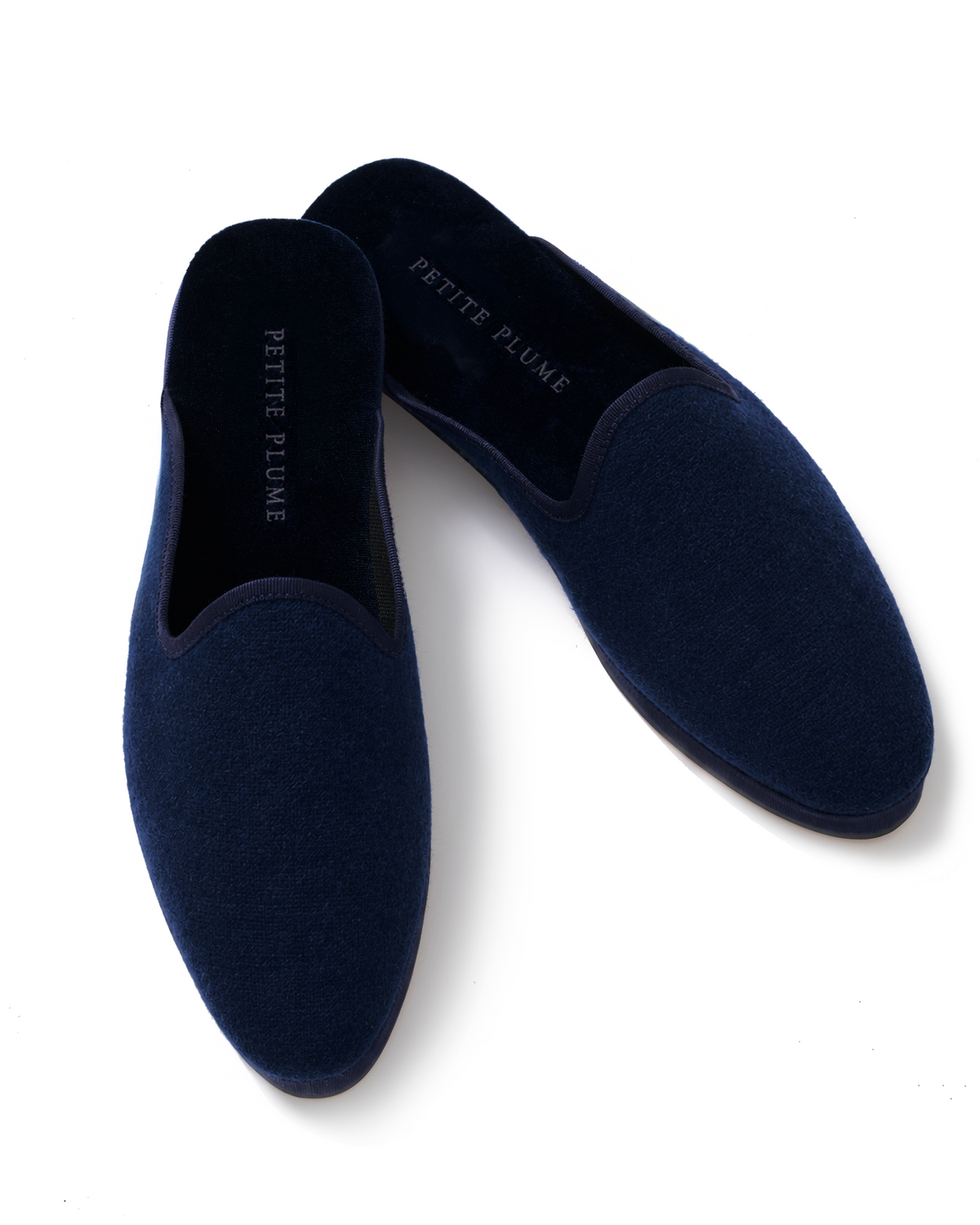 A pair of Women’s Slippers in Navy made of velvet with PETITE PLUME written in white on the insoles, shown on a white background.