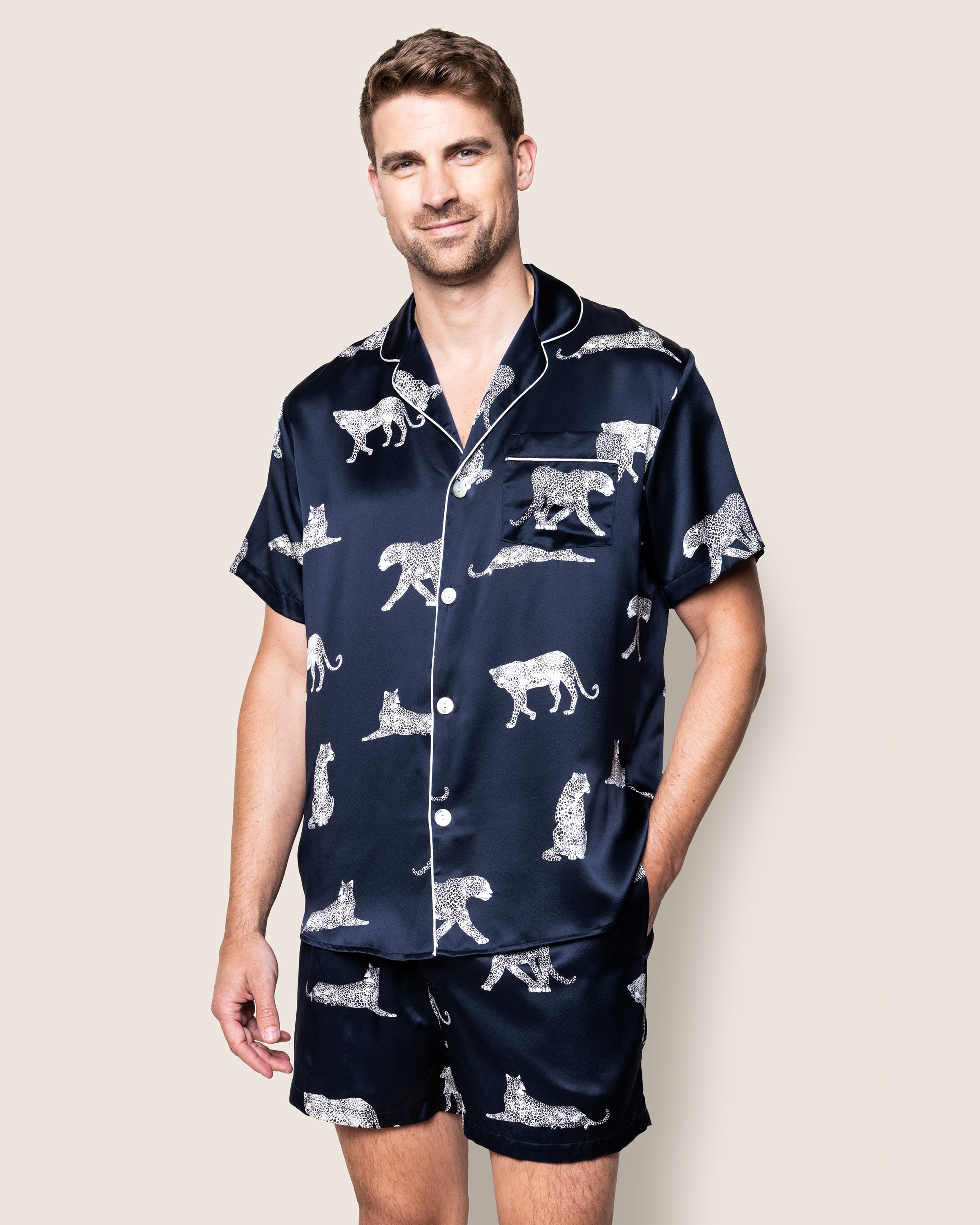 A man smiles in the Mens Silk Pajama Short Set in Panthère de Nuit by Petite Plume, featuring a matching navy hypoallergenic Mulberry silk with white leopard prints, including a button-up shirt and shorts.
