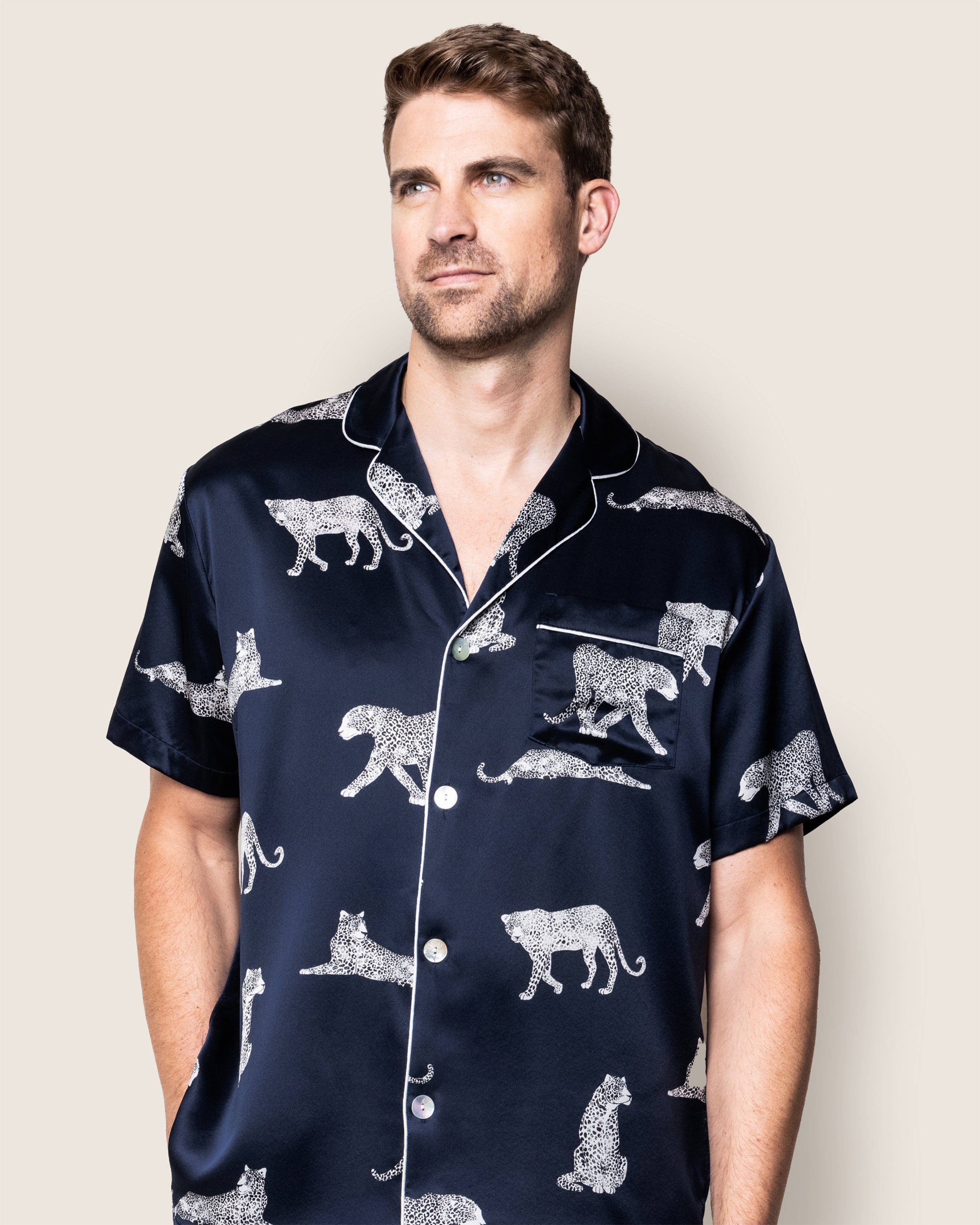 A man wears the Petite Plume Mens Silk Pajama Short Set in Panthère de Nuit, featuring a dark background with white leopard prints. Crafted from Mulberry silk, this luxurious sleepwear embodies elegance as he stands against a plain backdrop, gazing slightly to the side with a neutral look.