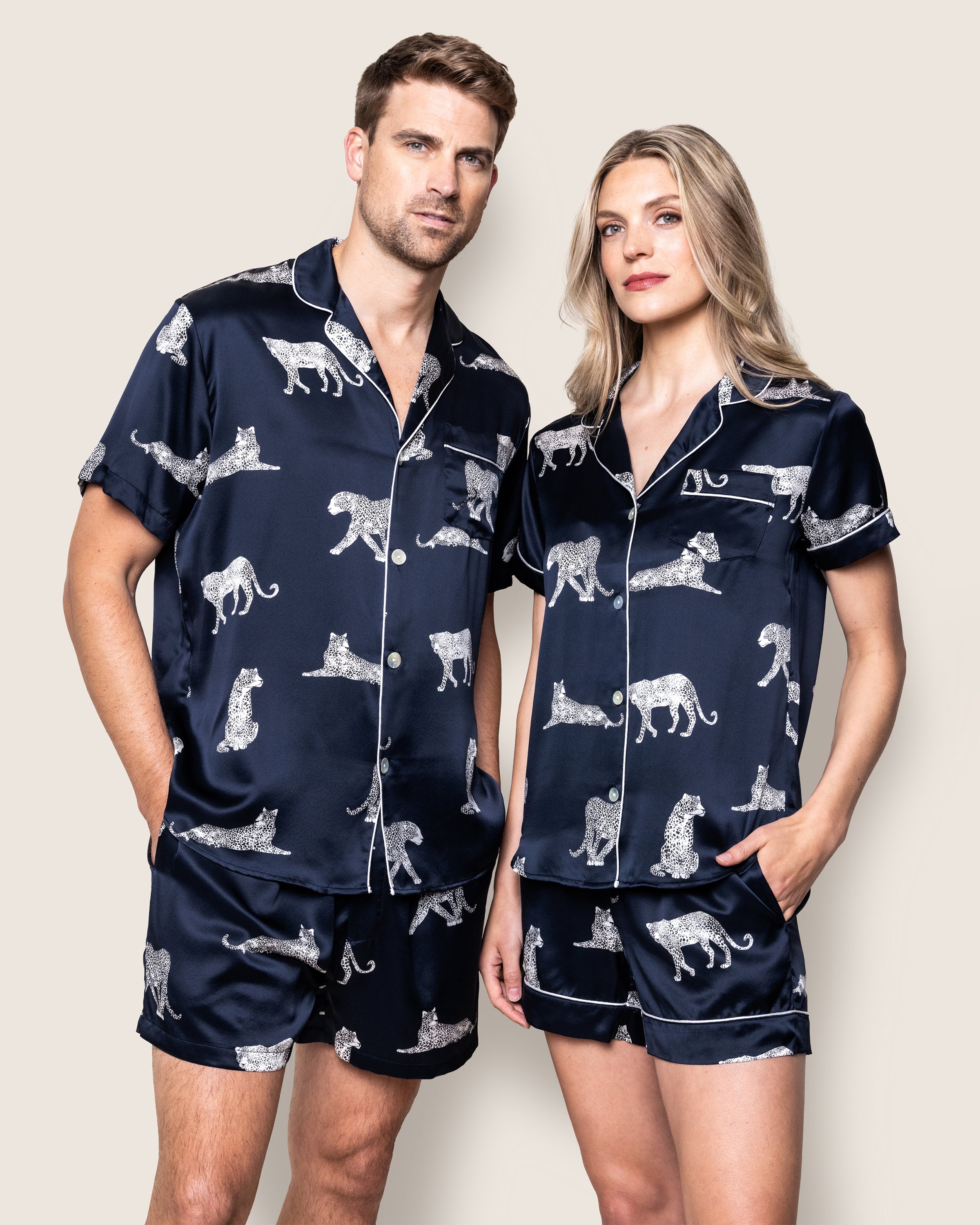A man and woman don matching navy blue Petite Plume silk pajamas, the Mens Silk Pajama Short Set in Panthère de Nuit. Made from hypoallergenic Mulberry silk with white animal prints, they stand side by side with hands in pockets against a plain backdrop, epitomizing luxury sleepwear.