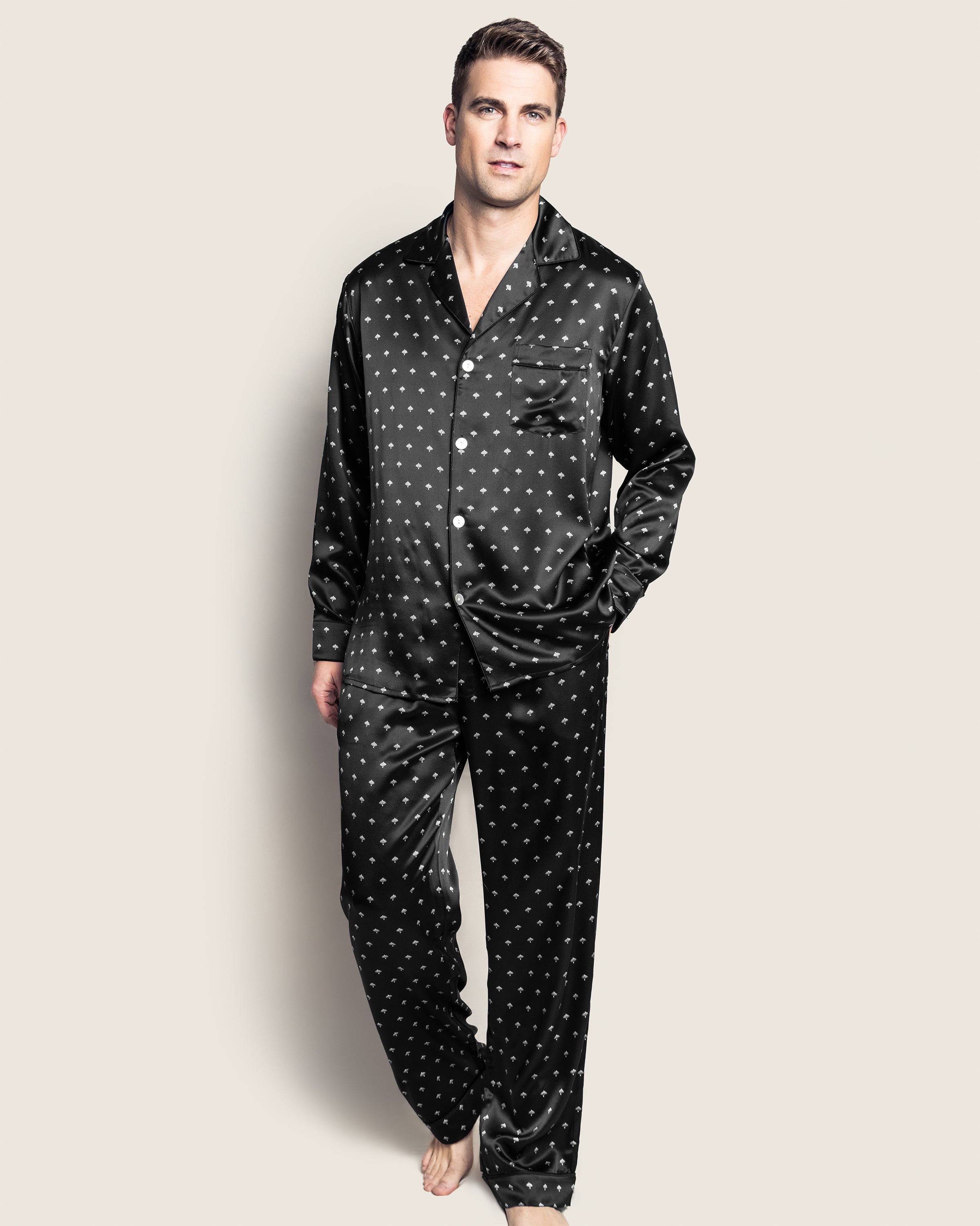 A man barefoot against a light background wears Petite Plumes Mens Silk Pajama Set in Black Art Nouveau. The elegant set, with a button-up shirt and matching pants, mirrors the luxury of mulberry silk. One hand in his pocket, he gazes slightly upward.