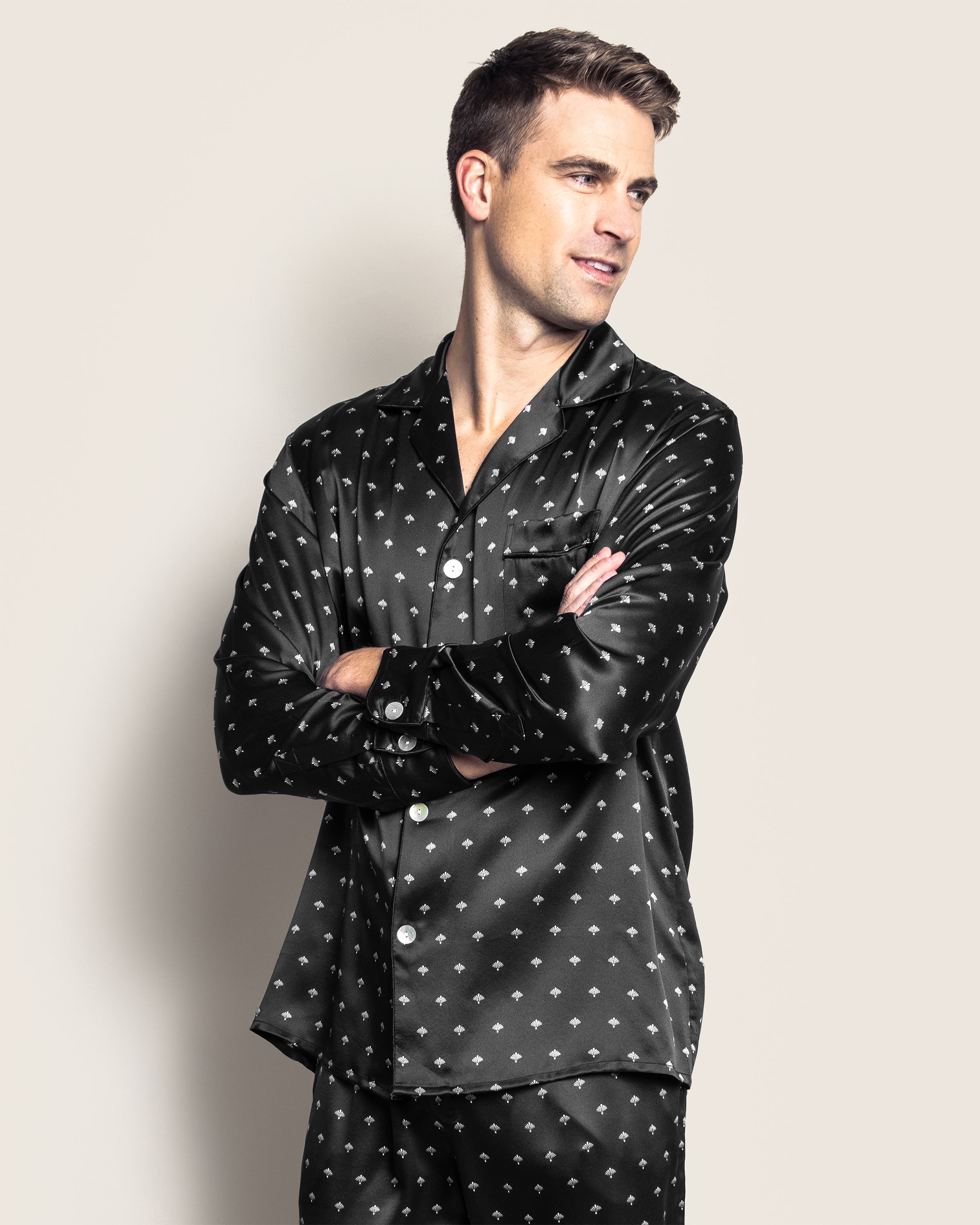 A man in a Mens Silk Pajama Set by Petite Plume, featuring a black Art Nouveau design, stands with arms crossed against a plain background, embodying luxury sleepwear.