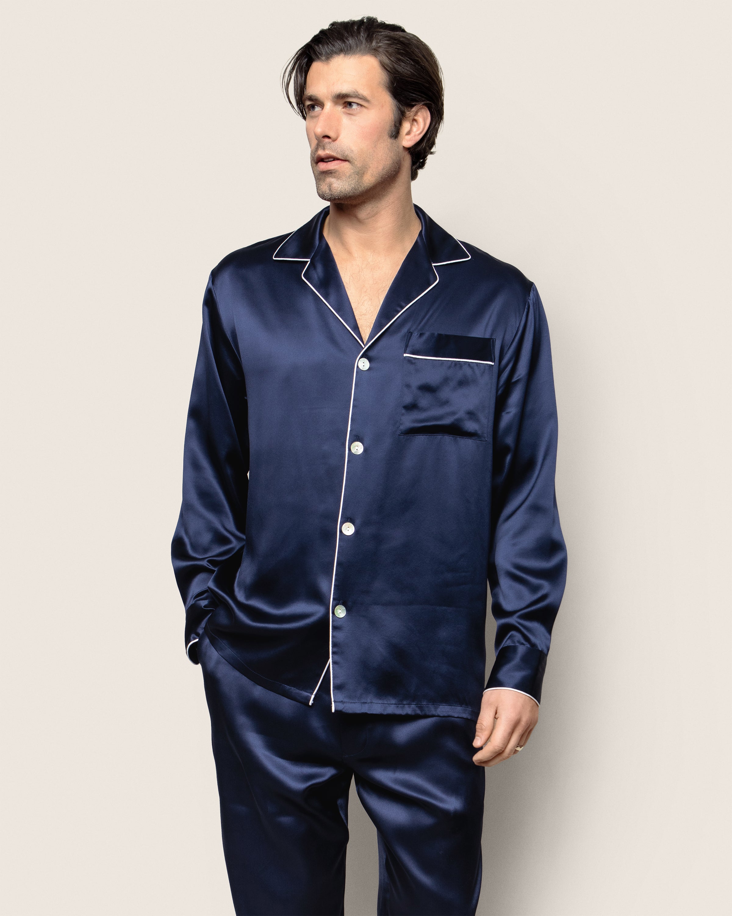 A man models Petite Plume’s Mens Silk Pajama Set in Navy, marked by white piping. He stands against a plain light background, gazing to the side with hands in pockets.