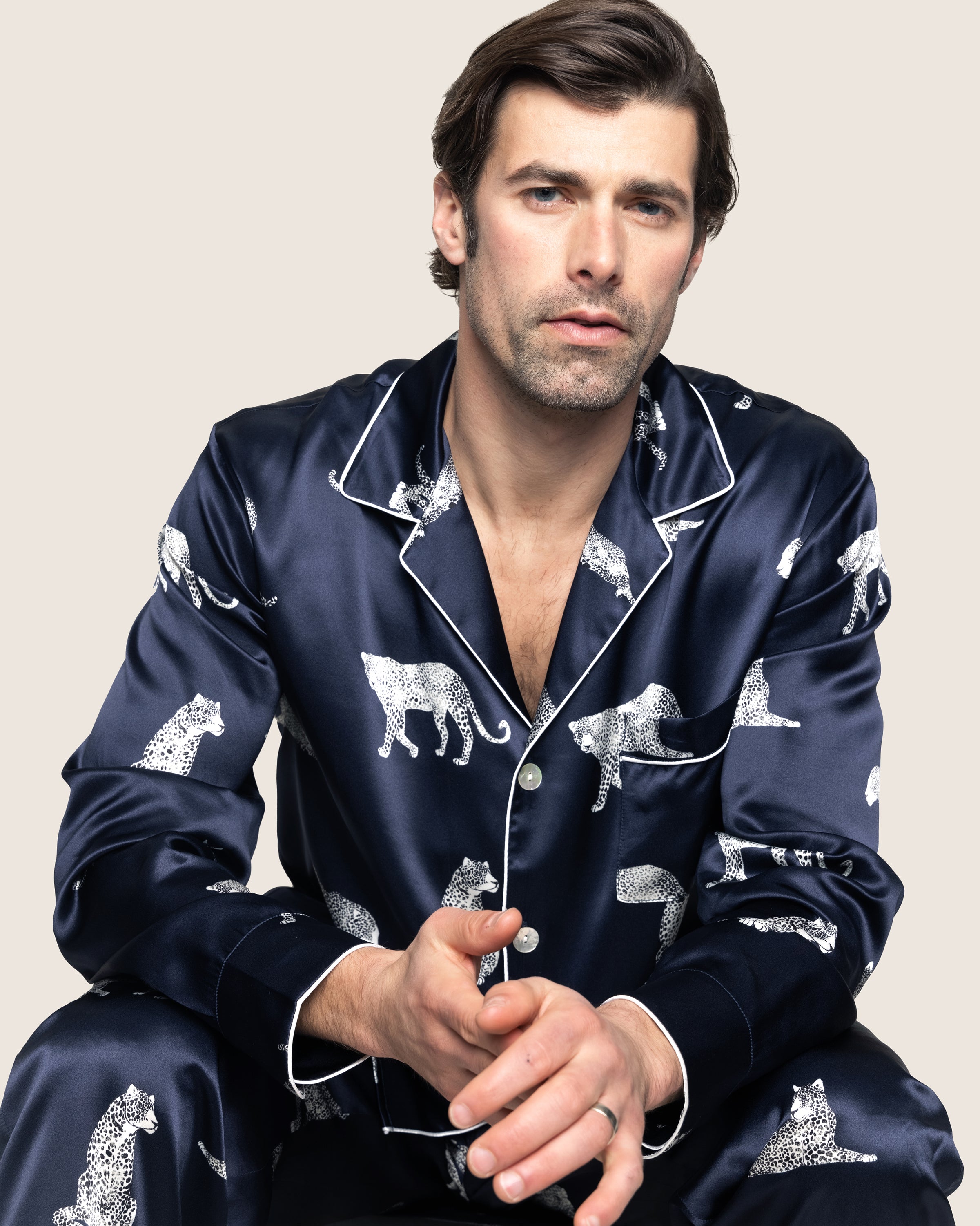 A man with dark hair and light facial hair sits looking forward, donning Petite Plumes Mens Silk Pajama Set in Panthère de Nuit, reminiscent of classic Hollywood style. The neutral solid background enhances the elegance of his silk sleepwear.