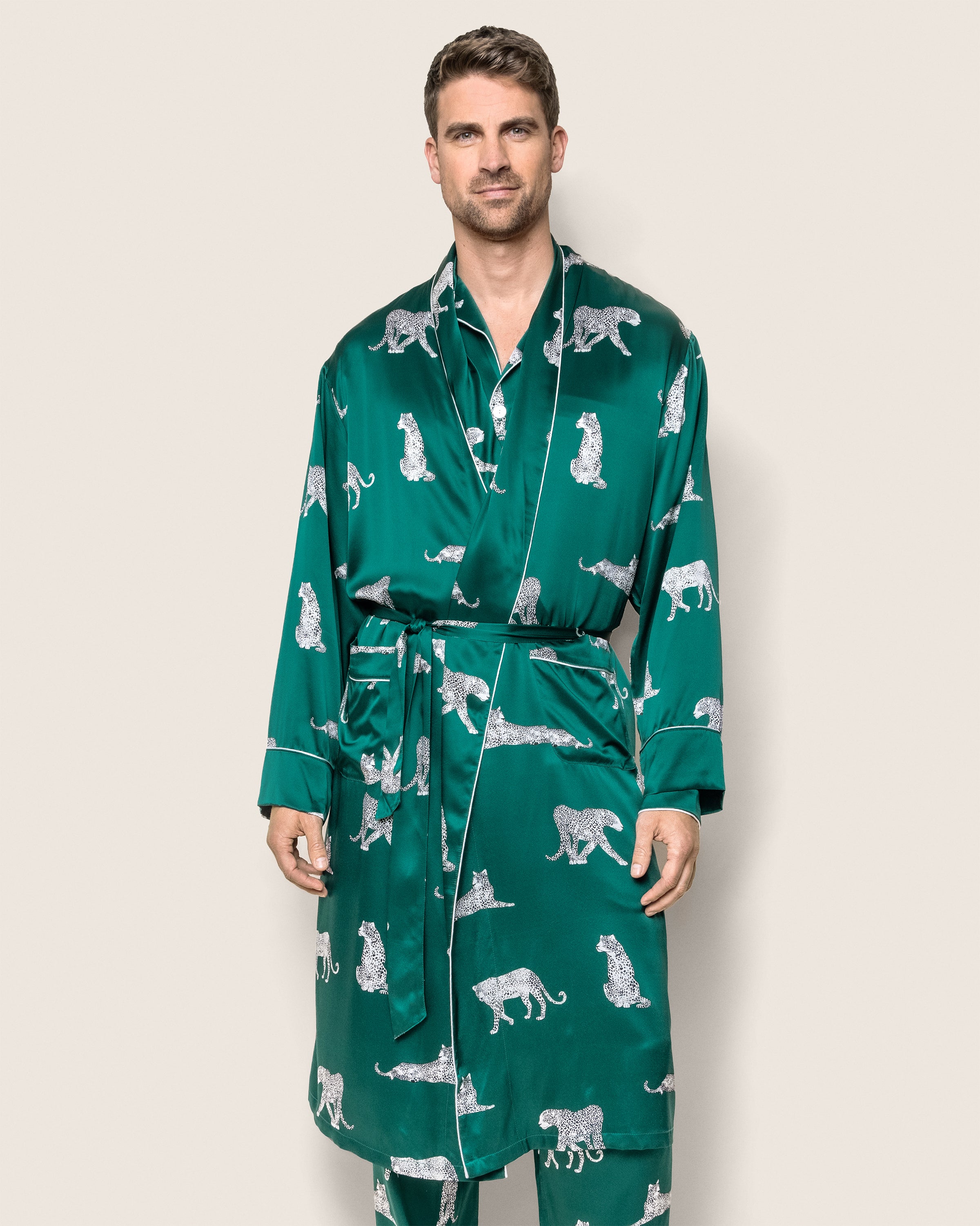 A man wears the Mens Silk Long Robe in Panthère de Luxe by Petite Plume, featuring a green Mulberry silk with white leopard patterns. The robe ties at the waist, adding style to his look against a plain backdrop.