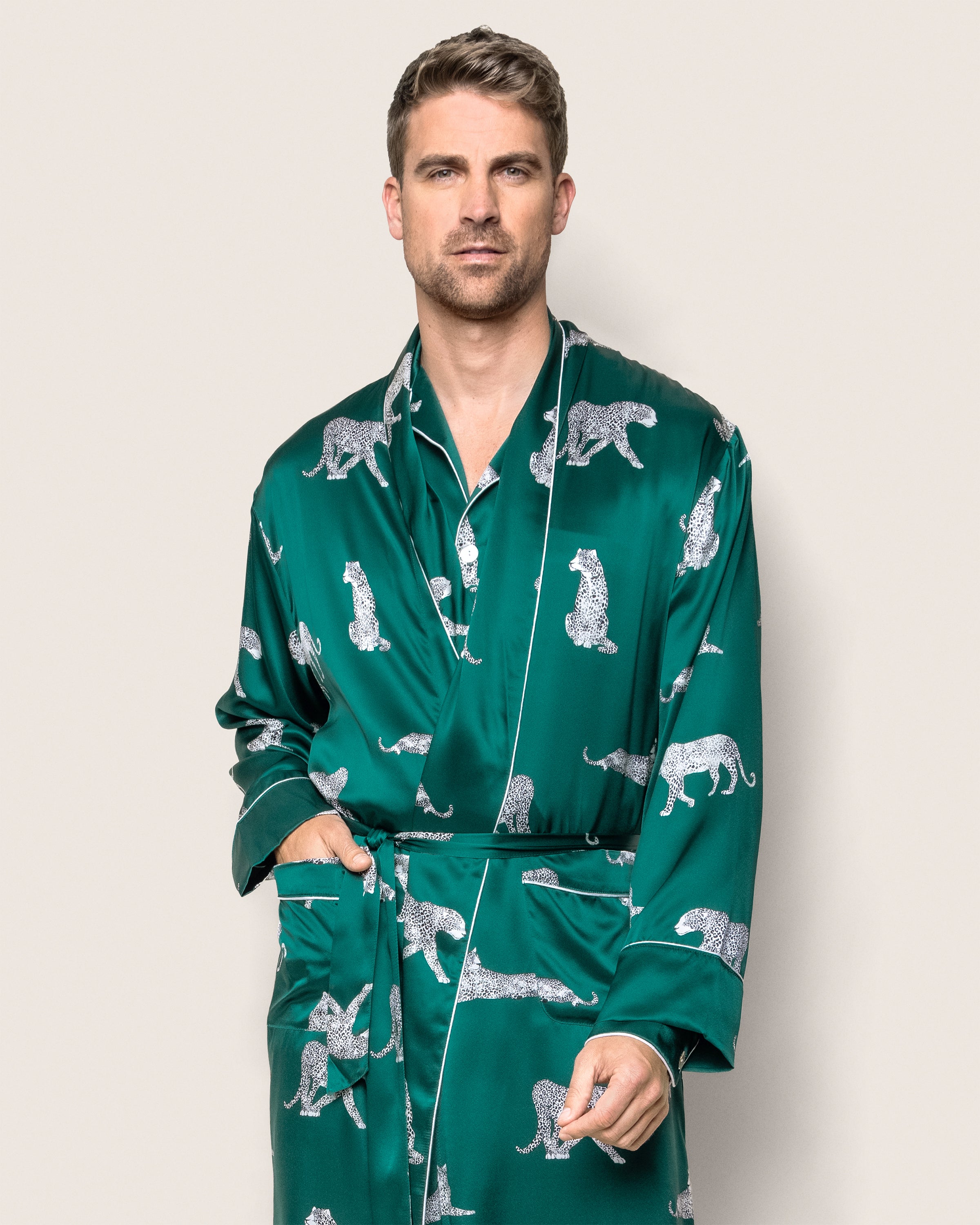 A man in a Mens Silk Long Robe in Panthère de Luxe from Petite Plume, featuring elegant green mulberry silk with white leopard prints, stands against a beige background. His styled short hair and relaxed posture, with one hand in a pocket, exude elegance and upscale sleepwear.