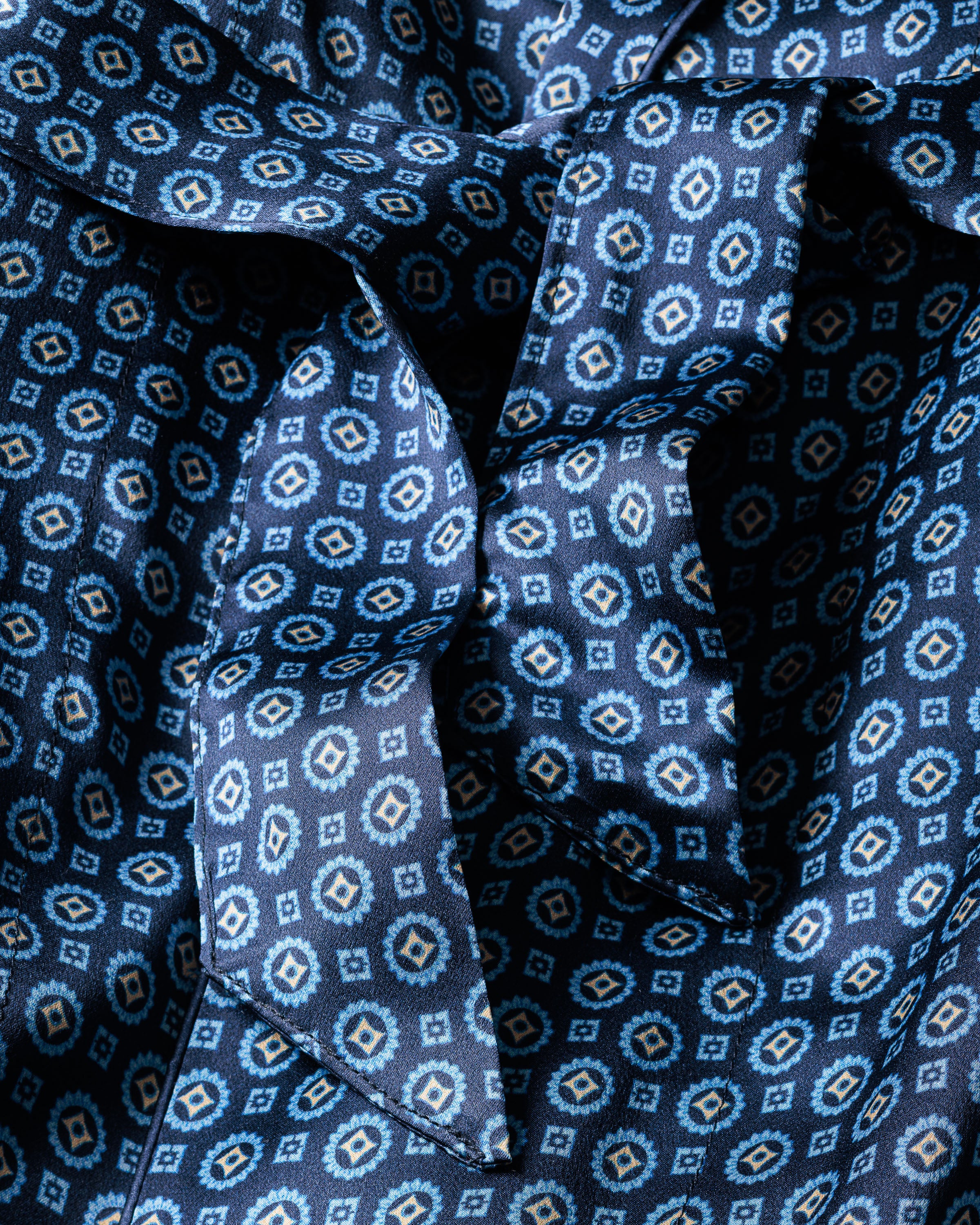 A close-up of the Mens Silk Robe in Midnight Foulard by Petite Plume reveals its blue fabric with white and light blue hexagonal patterns. Reminiscent of luxurious Mulberry silk, its textured and slightly crumpled design under soft lighting makes it perfect for breathable sleepwear.