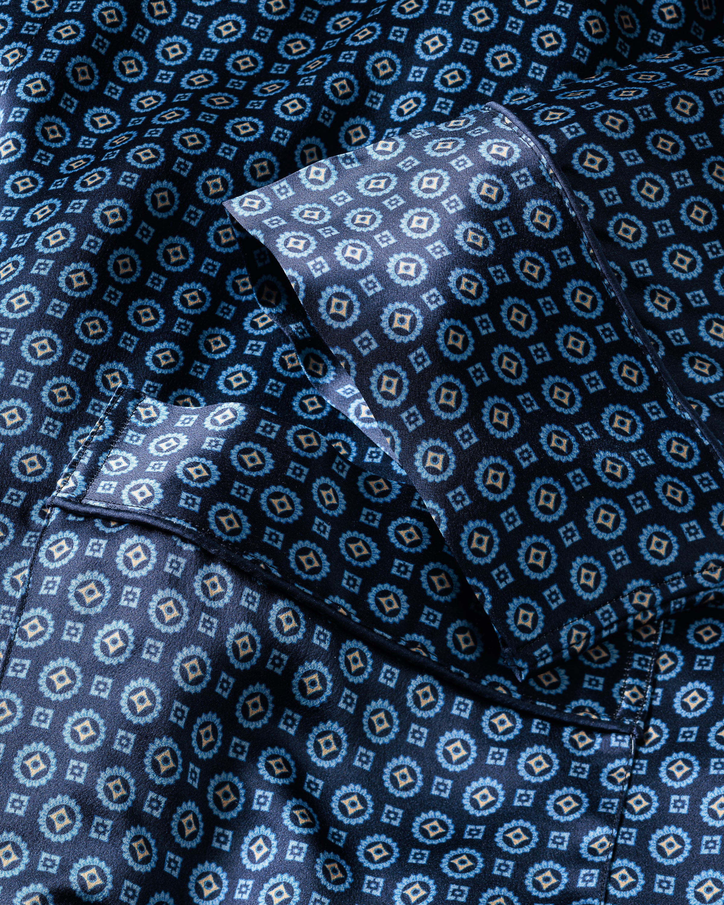 The Mens Silk Robe in Midnight Foulard by Petite Plume features a silky, breathable fabric with a geometric pattern of hexagons and small diamonds in blue and light tan, offering a textured, shiny appearance perfect for luxurious sleepwear.